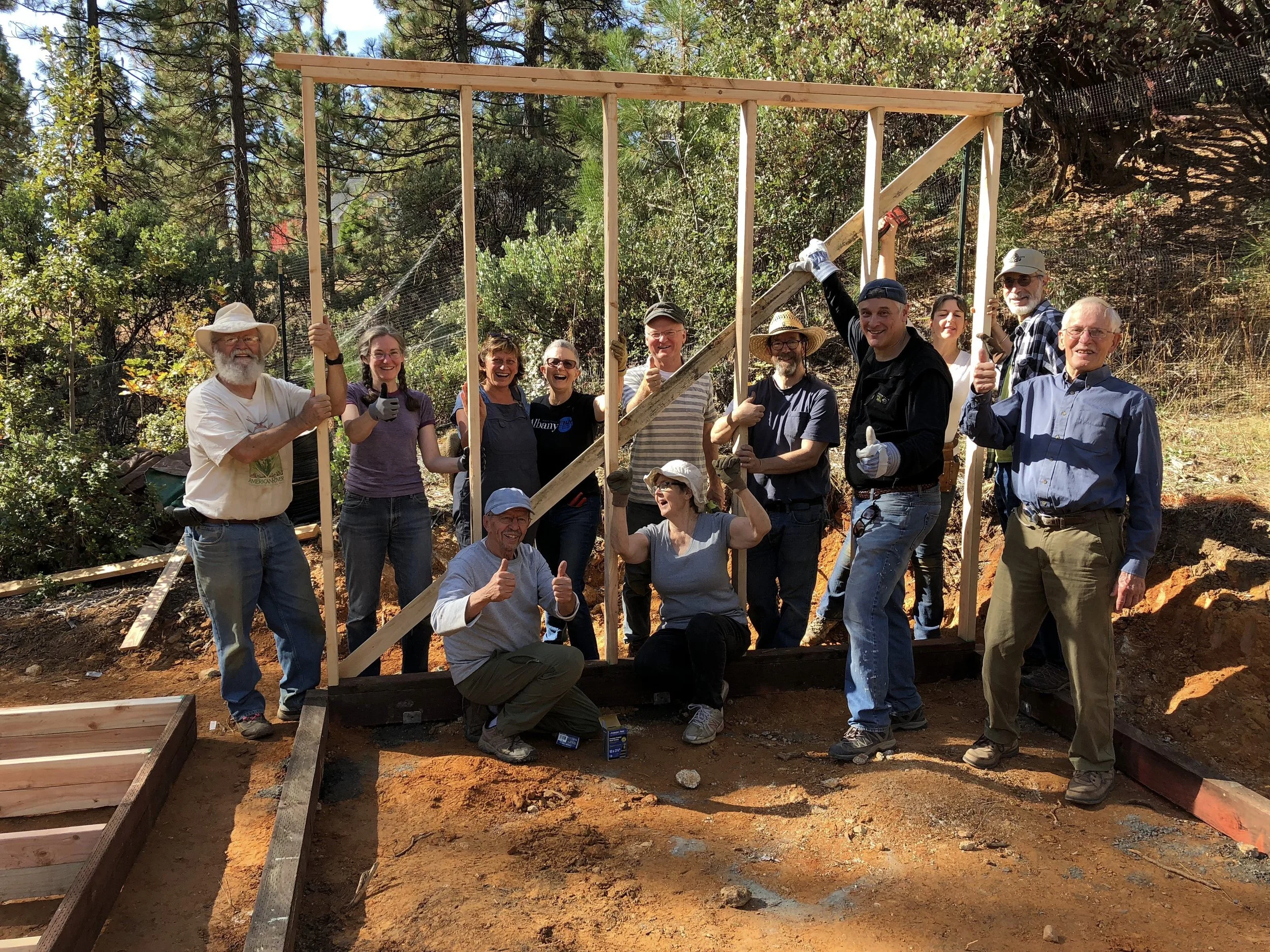 A group of people, including men and women of various ages, standing outdoors on a construction site surrounded by trees and shrubs. They are smiling, giving thumbs up, and holding wooden framing pieces for a building under construction.