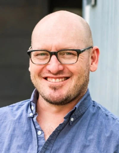 A man with a bald head, glasses, and a beard smiling outdoors, wearing a blue button-up shirt.