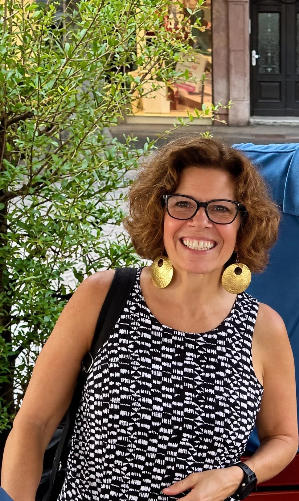 Smiling woman with curly hair in a colorful dress sitting outside at a table with string lights in the background.