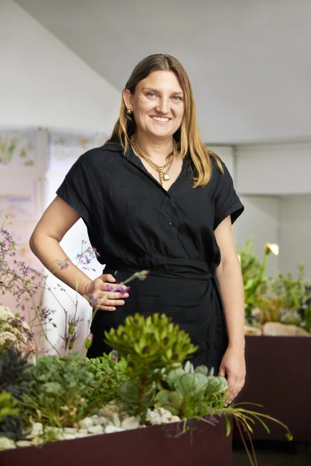 A woman with shoulder-length brown hair, wearing a black dress, gold jewelry, and smiling, standing next to a table with various succulents and plants in an indoor setting.