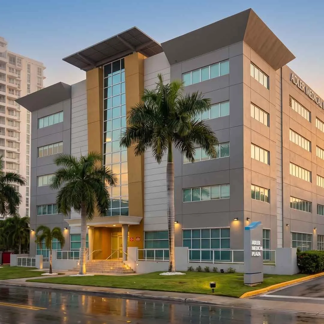 Edificio de clínica o hospital con palmeras y señalética que dice 'Adler Medical Plaza' en un entorno urbano con cielo al atardecer.