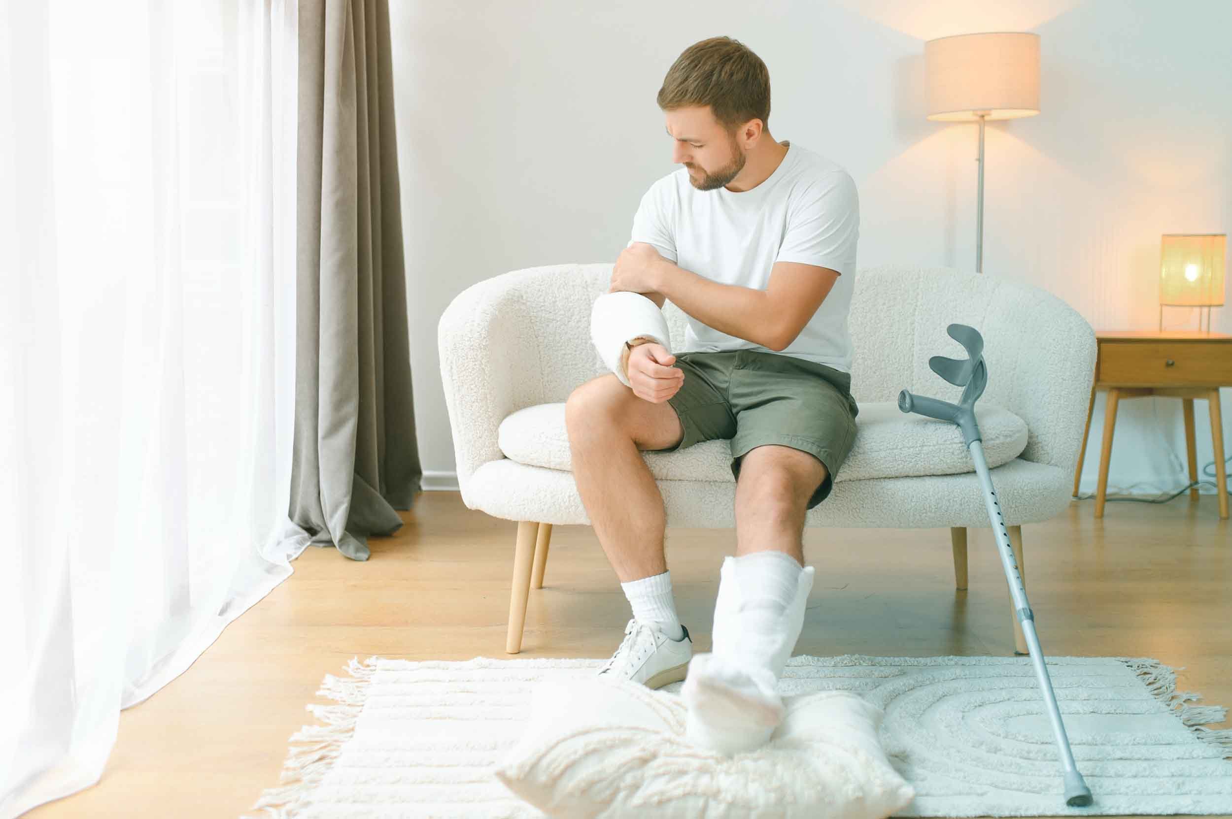 Joven sentado en un sillón, sosteniéndose la muñeca izquierda con expresión de dolor, junto a una muleta apoyada en el suelo, en una habitación con luz natural y moderna decoración.