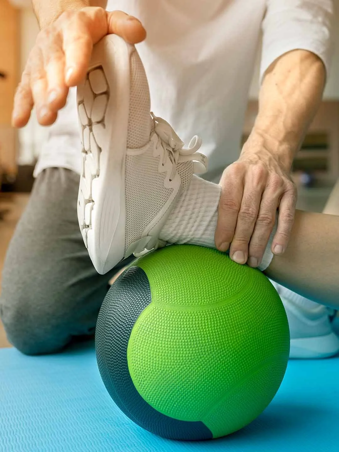 Persona mayor realizando ejercicios de rehabilitación con una pelota de masaje verde, levantando la pierna de otra persona en un ambiente interior.