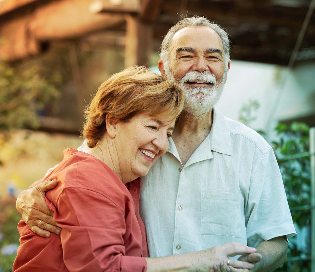 Una pareja de ancianos sonriendo y abrazándose en un entorno al aire libre, con vegetación de fondo.