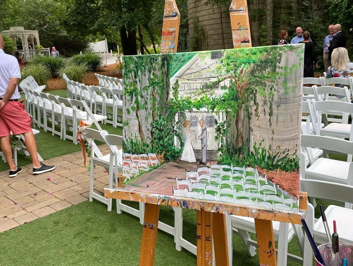 Painting of a wedding ceremony scene with a bride and groom holding hands, surrounded by greenery, displayed on an easel at an outdoor wedding setup.