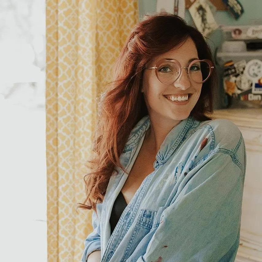 A young woman with long red hair, wearing glasses and a blue denim shirt, smiling while standing indoors near a yellow patterned curtain.