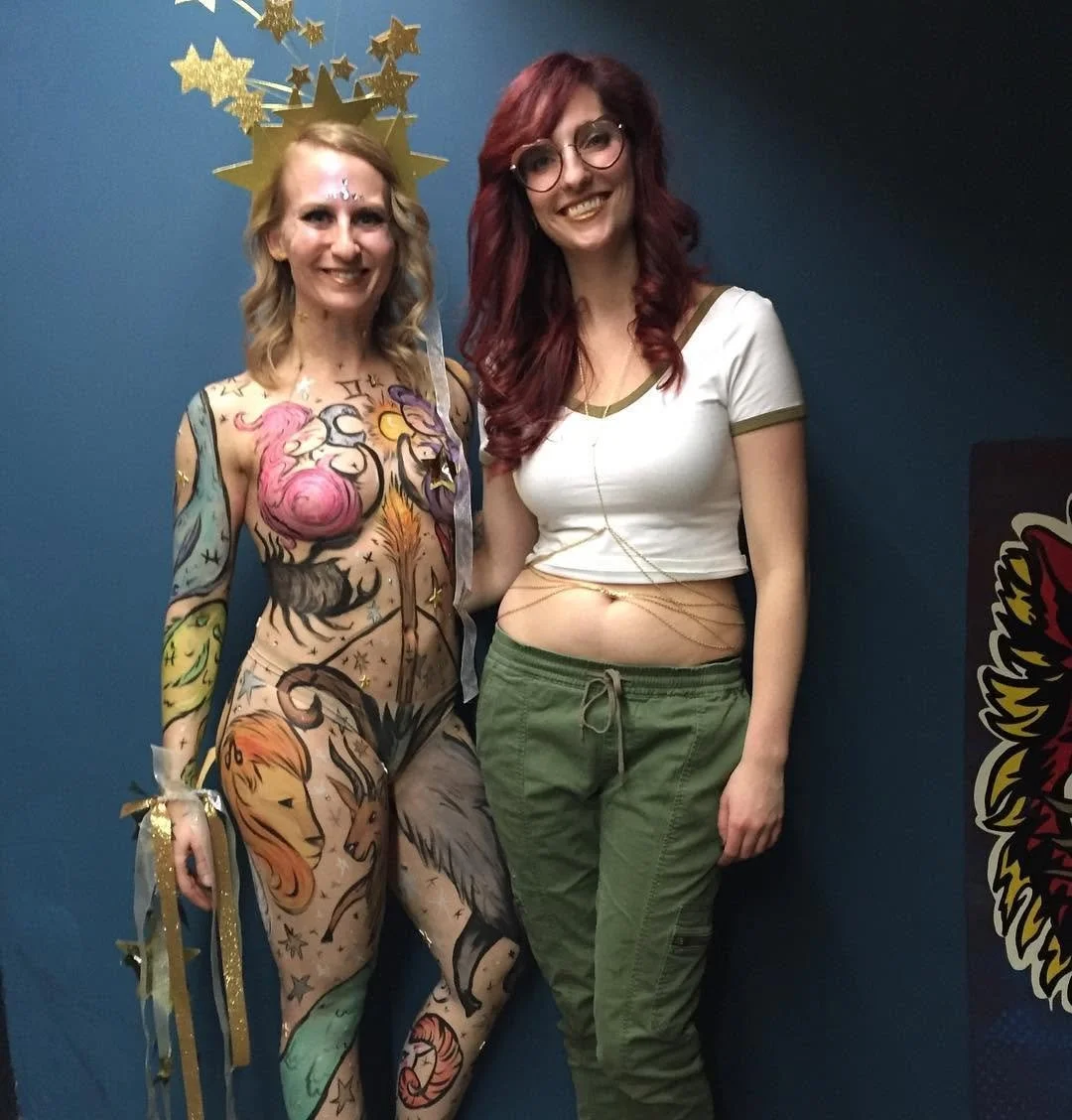 Two women standing together against a blue wall, one in full body paint with colorful zodiac and star designs and a gold star crown, the other wearing glasses, a white and green t-shirt, and green pants, smiling at the camera.