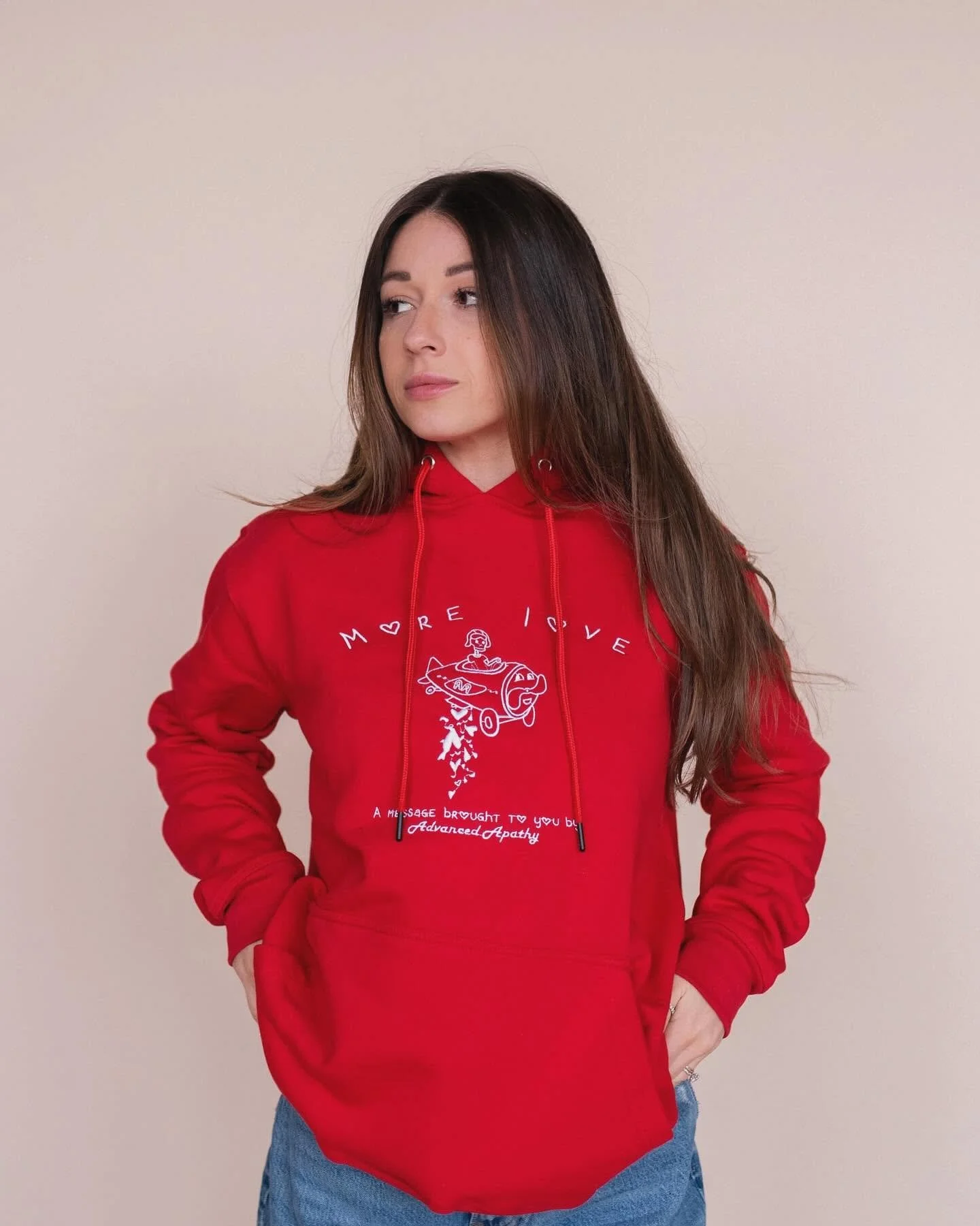 Young woman with long brown hair wearing a red hoodie and blue jeans standing against a beige wall.