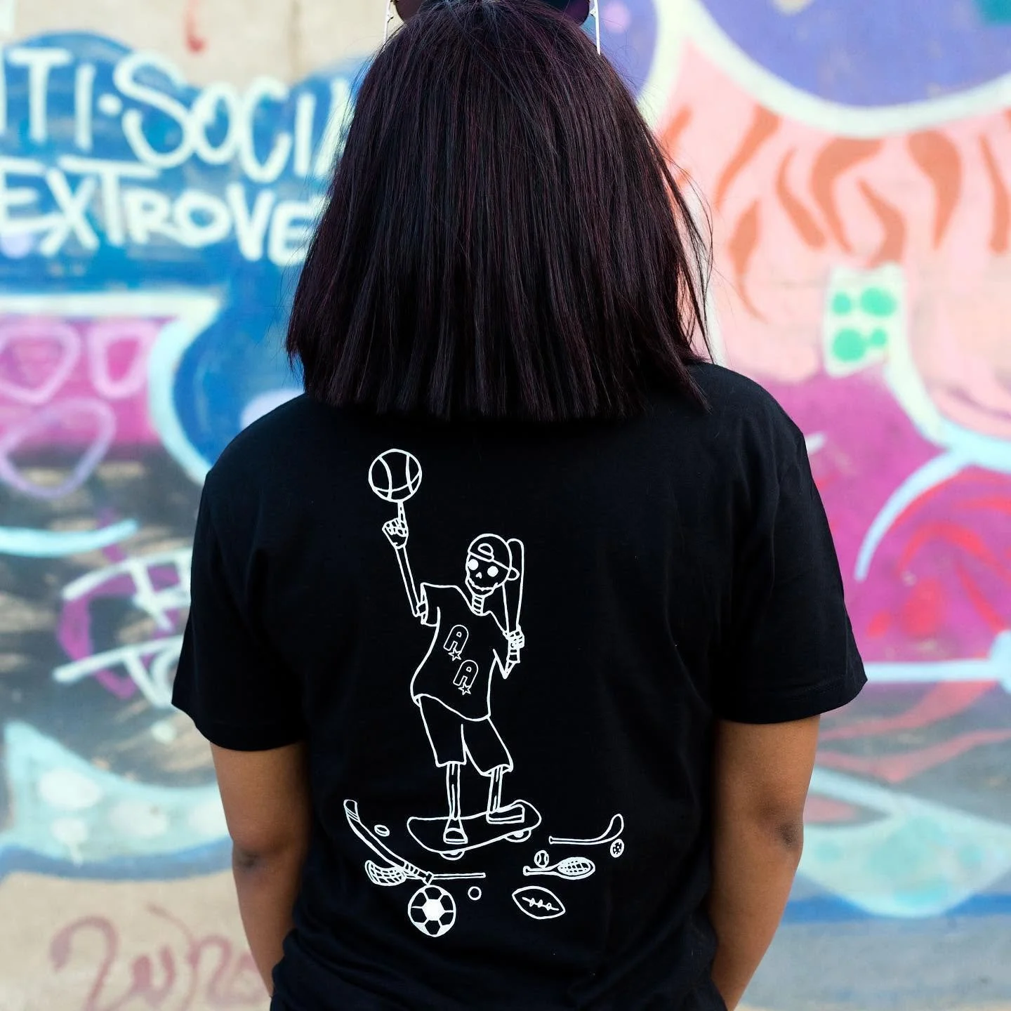 Person with dark hair wearing a black T-shirt with a white line drawing of a skeleton skateboarding, holding a basketball, with sporting goods around the figure illustrated on the shirt, standing in front of a colorful graffiti wall.