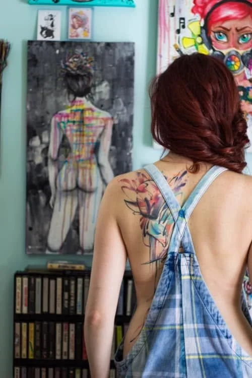 A woman with reddish hair and tattoos on her back, wearing plaid blue overalls, standing in an art studio or gallery with colorful artwork on the walls.