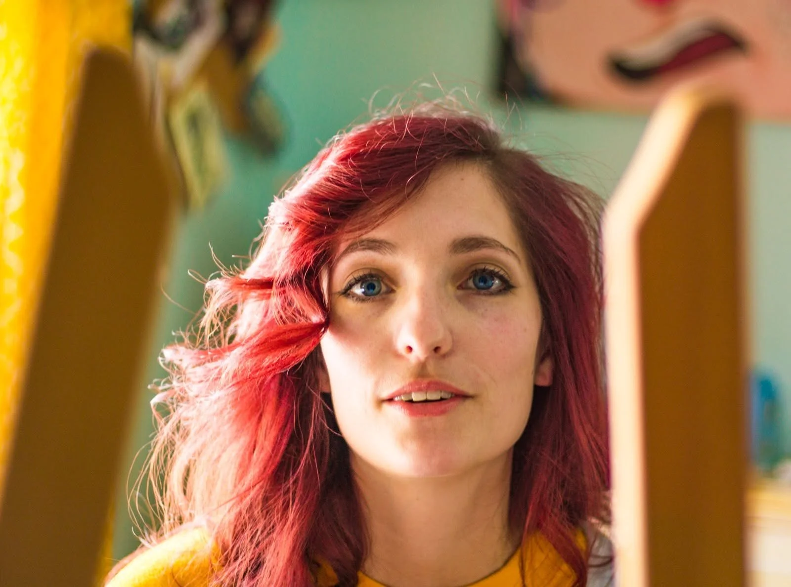 A young woman with bright red hair and blue eyes looking through the top of an easle with blurred background including a colorful painting.