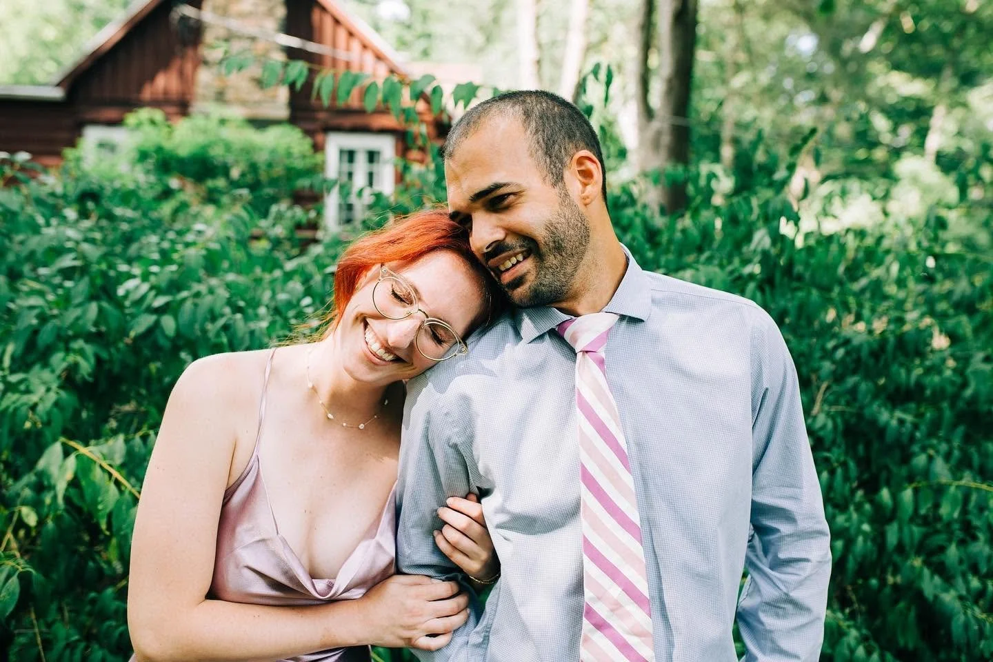 A couple smiling and cuddling outdoors with a house in the background.