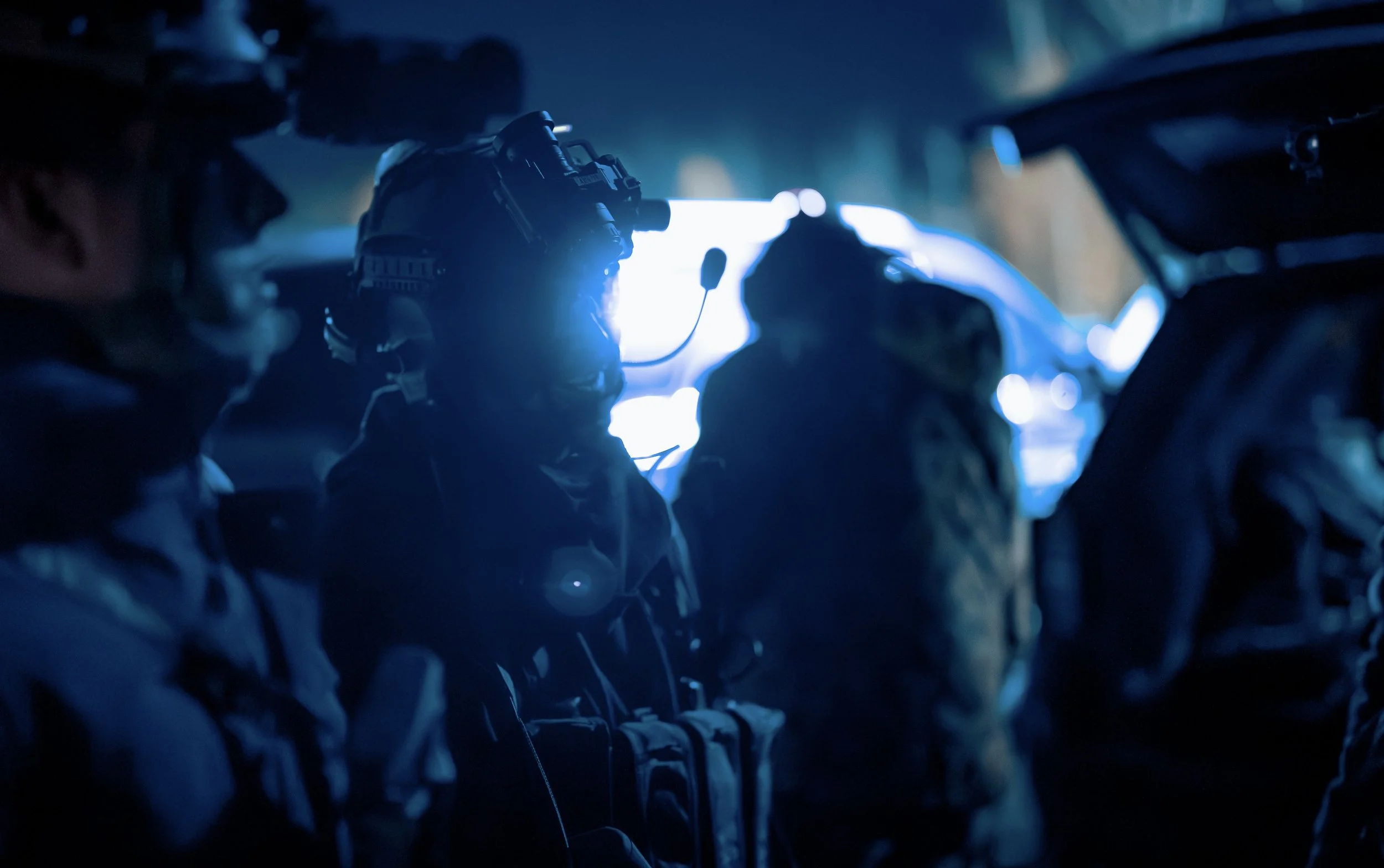 Silhouettes of people wearing headsets inside an aircraft cockpit or control room with illuminated screens and equipment, viewed from behind in a dark environment.