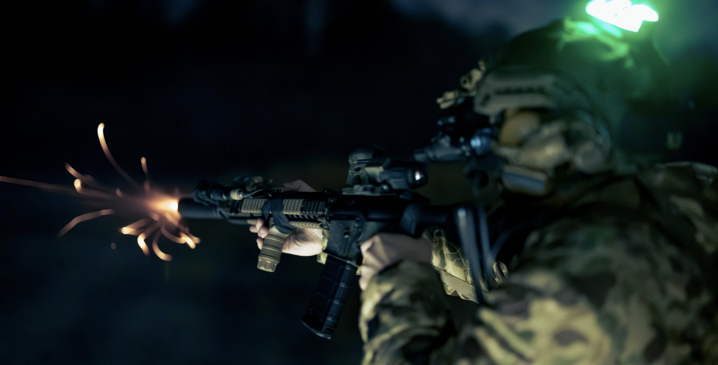 A soldier in camouflage gear aiming a rifle during nighttime, with sparks and flashes coming from the firearm.
