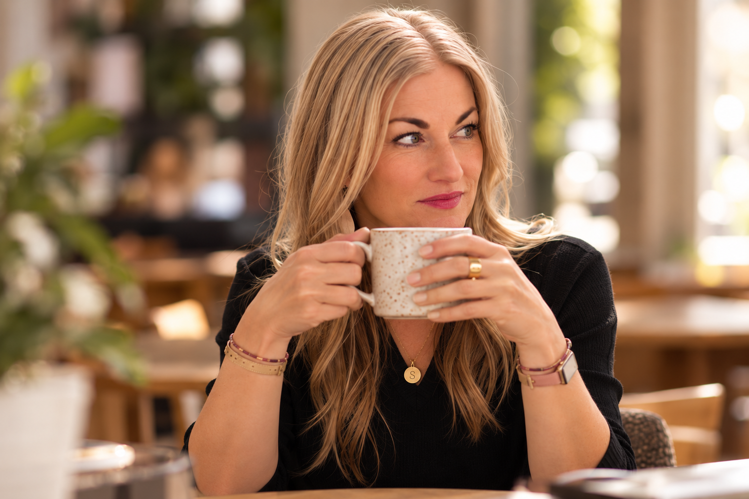 A blonde woman in a black top sitting in a cozy coffee shop, holding a speckled coffee mug and looking to the side with a slight smile, wearing a gold necklace with an 'S' pendant, a watch, and bracelets.