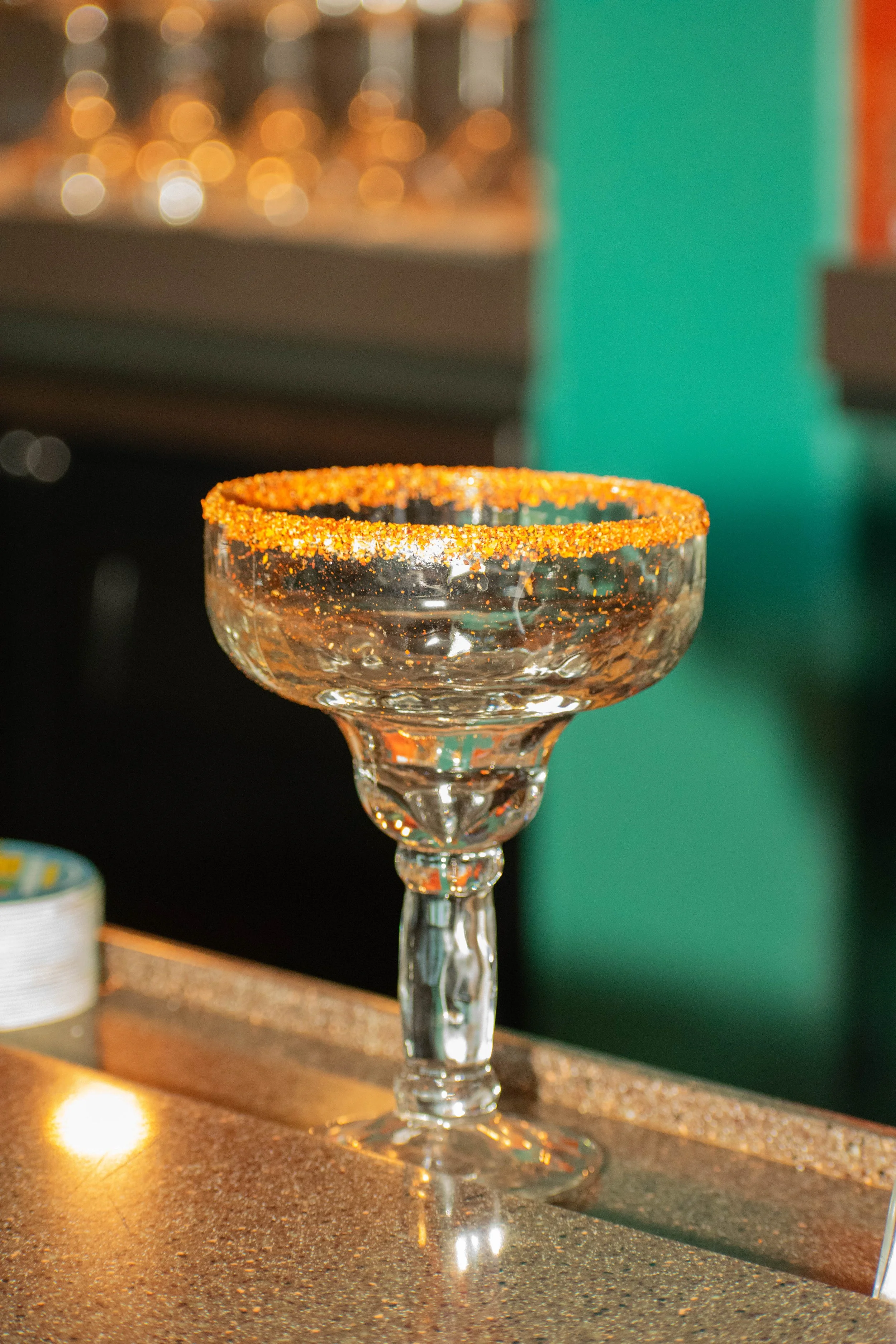 A clear glass margarita cocktail glass with an orange chili salt rim, placed on a glittery gold surface, with a blurred background.