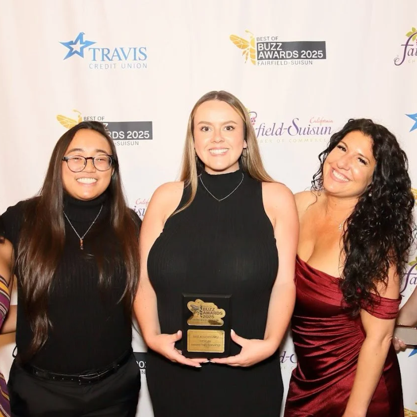 Three women smiling at an awards ceremony with a backdrop displaying logos for Travis Credit Union, Buzz Awards 2025, and Fairfield-Suisun, one woman in a black dress holding an award plaque.