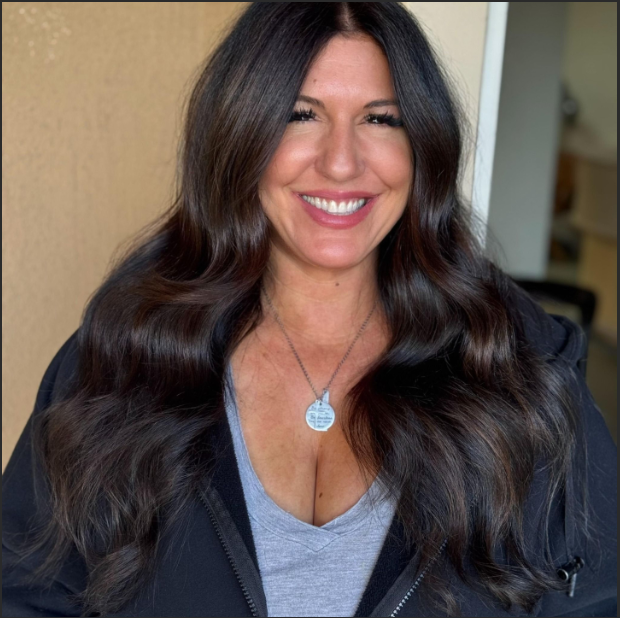 Smiling woman with long, wavy dark hair wearing a grey top and black jacket, posing indoors.