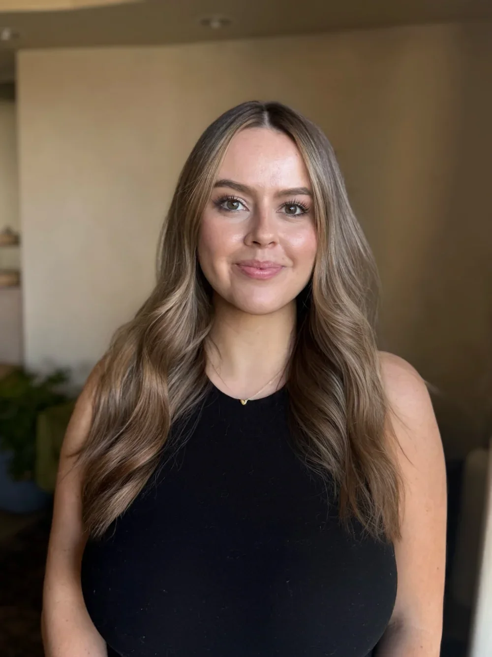 A young woman with long wavy blonde hair, wearing a black sleeveless top, smiling softly at the camera, in a room with a neutral background.
