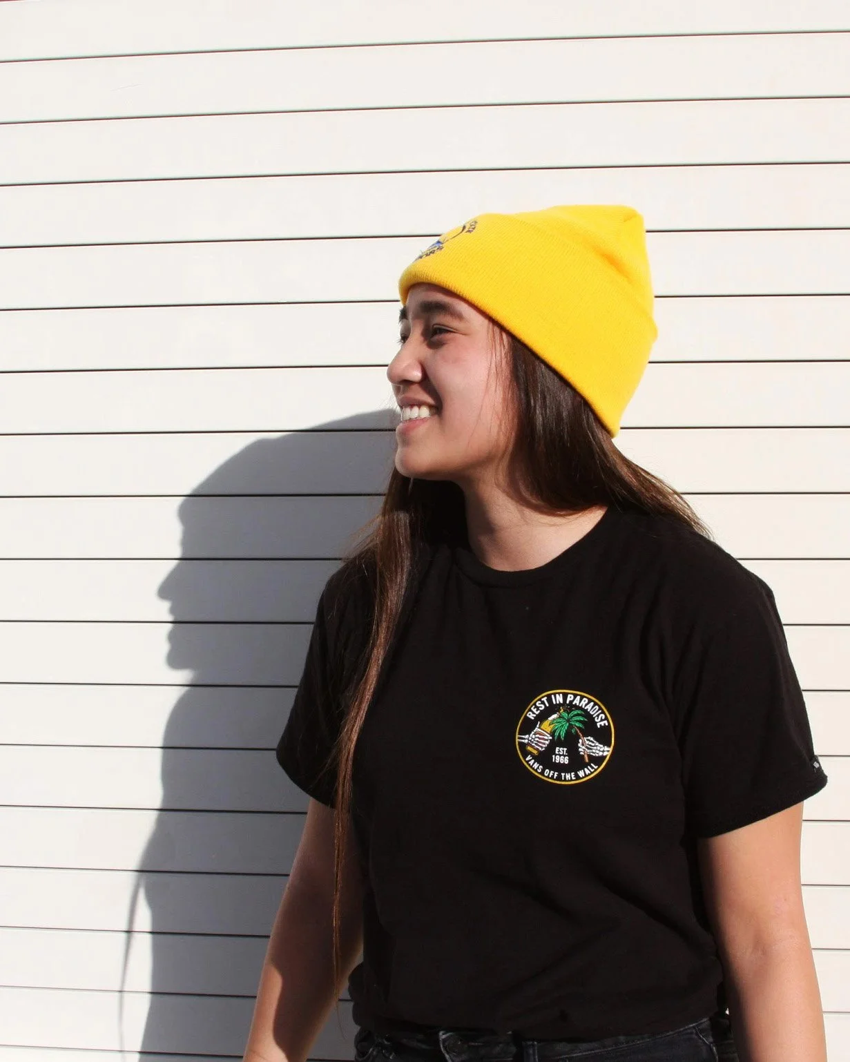 Young woman wearing a yellow beanie and black t-shirt with a logo, standing against a white paneled wall with shadows.