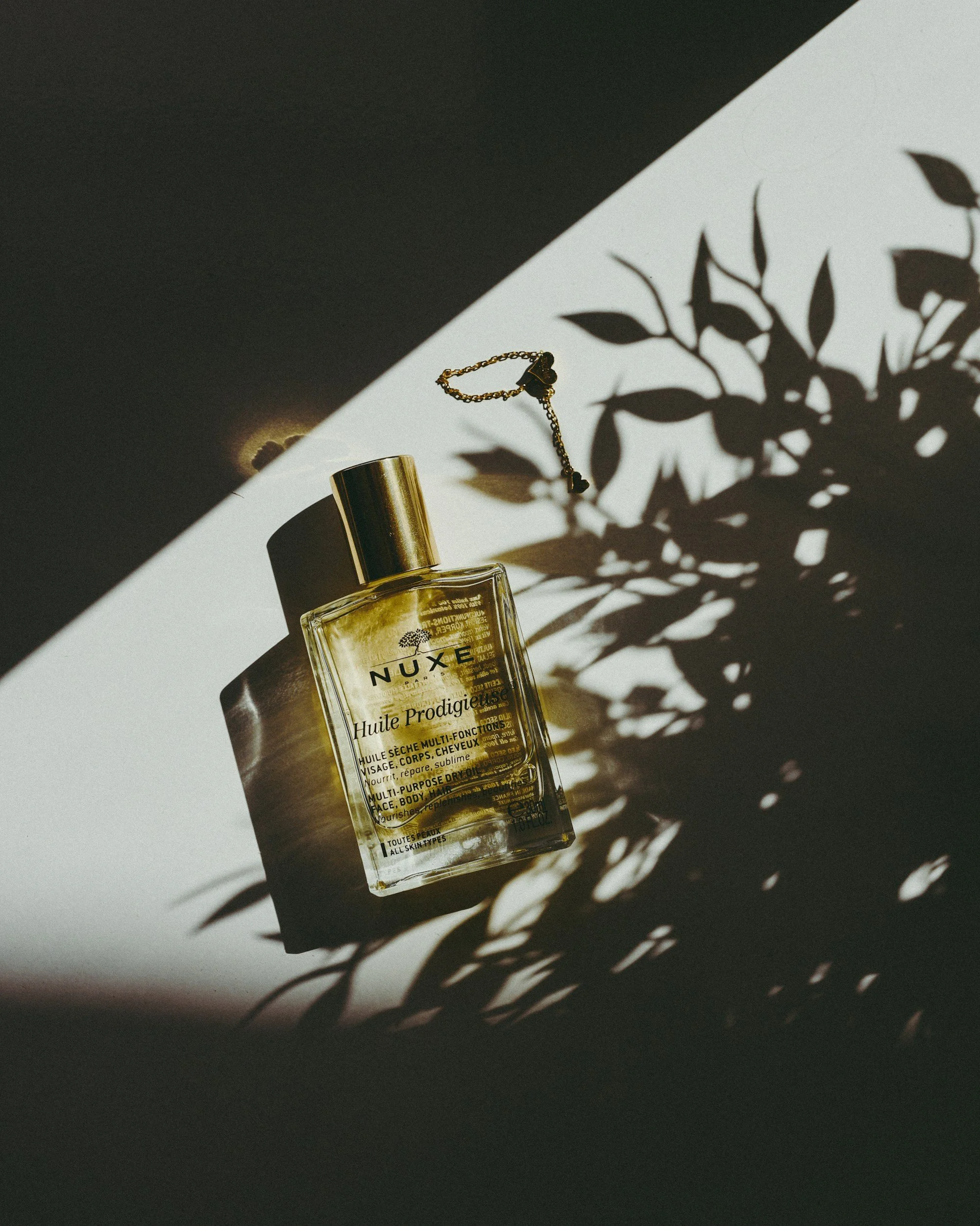 A clear glass bottle of Nuxe Huile Prodigieuse dry oil with a gold cap, casting a shadow of leaves on a two-tone background.
