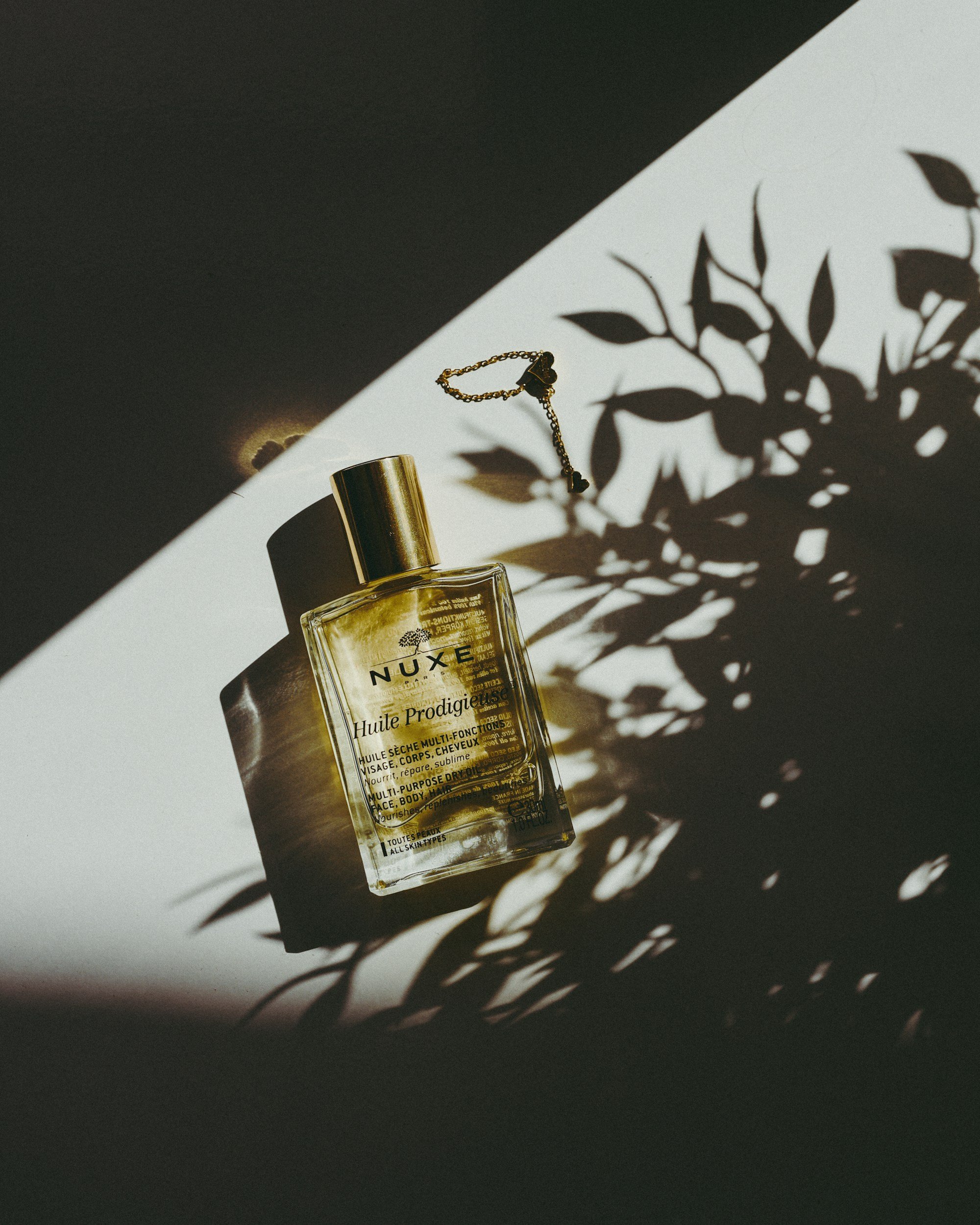 A bottle of NUXE oil with a gold cap and a gold wristwatch with a heart-shaped charm, casting shadows of a plant on a white and dark background.