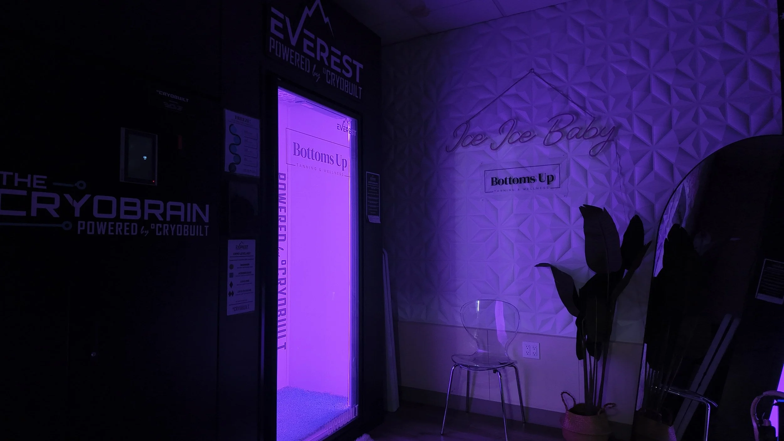 Inside a wellness center with purple lighting, there's an elevator with branding reading 'Everest Powered by Cryobuilt' and 'Bottoms Up Tanning & Wellness.' A neon sign on the wall says 'Ice Baby.' There are two clear chairs, a potted plant, and a small table.