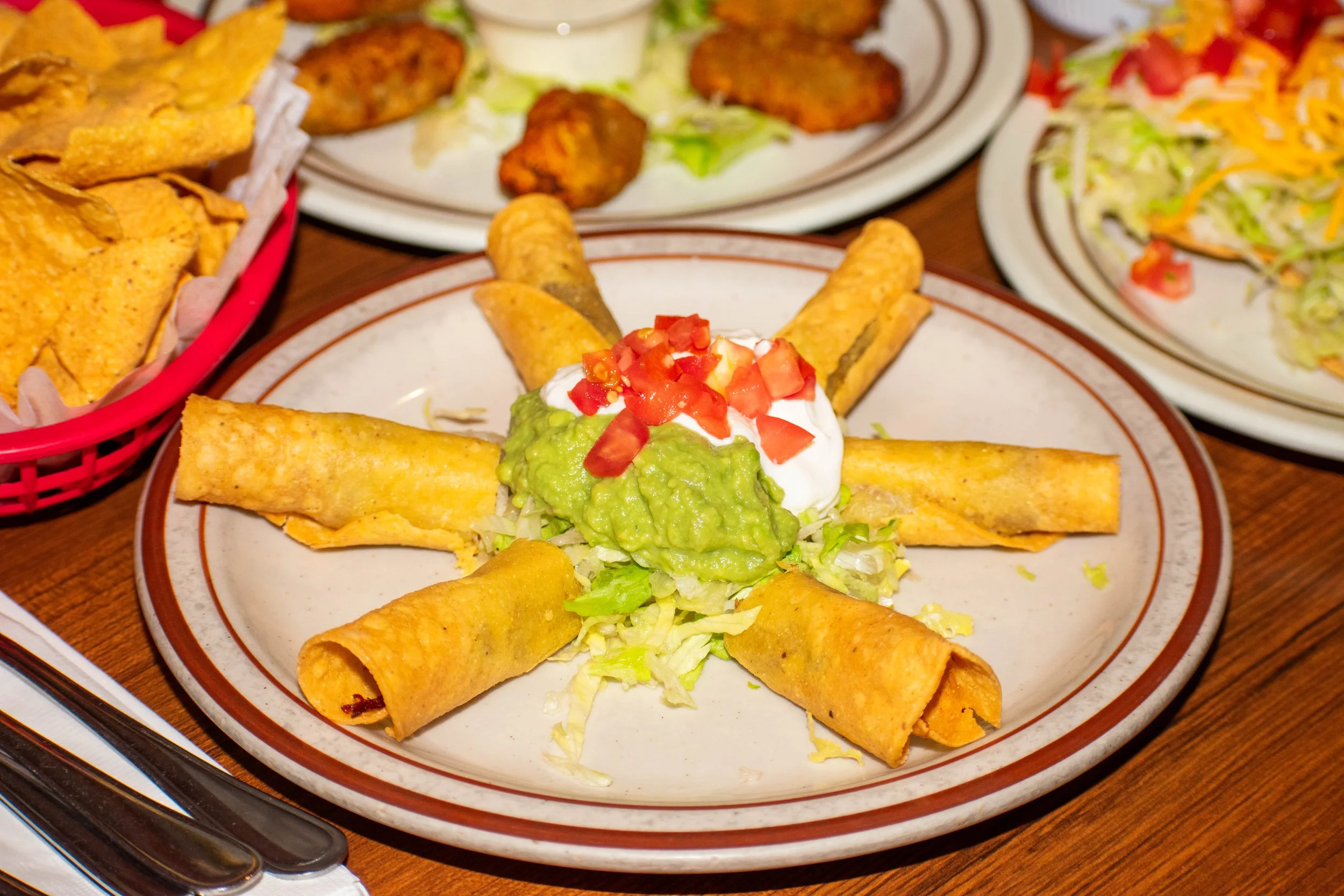 Plate of Mexican food with tacos filled with lettuce, guacamole, sour cream, and diced tomatoes, surrounded by other plates of chicken wings, salad, and chips.