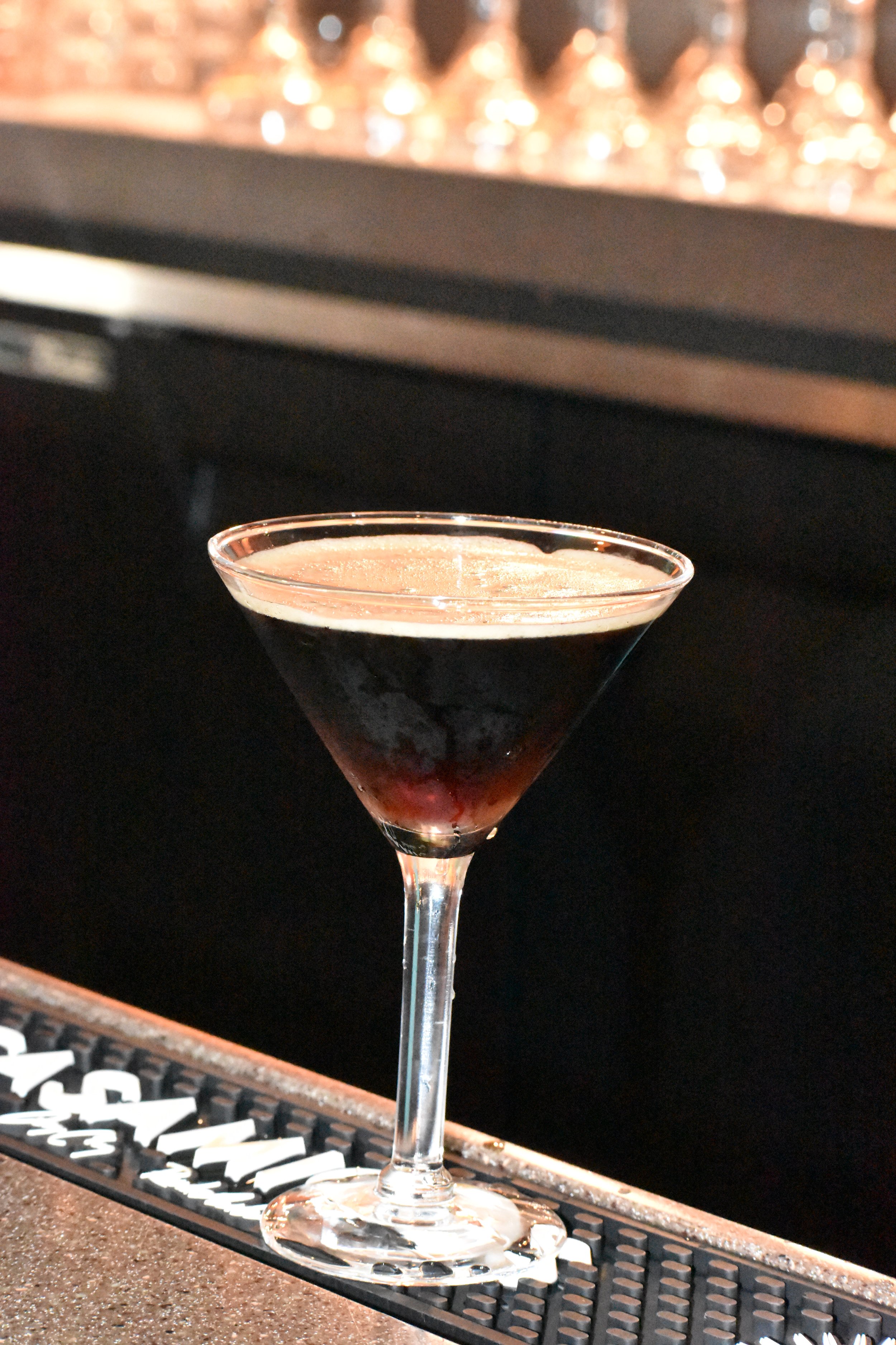 A cocktail glass filled with a dark red liquid, sitting on a bar counter with a black rubber bar mat and a lit bar in the background.