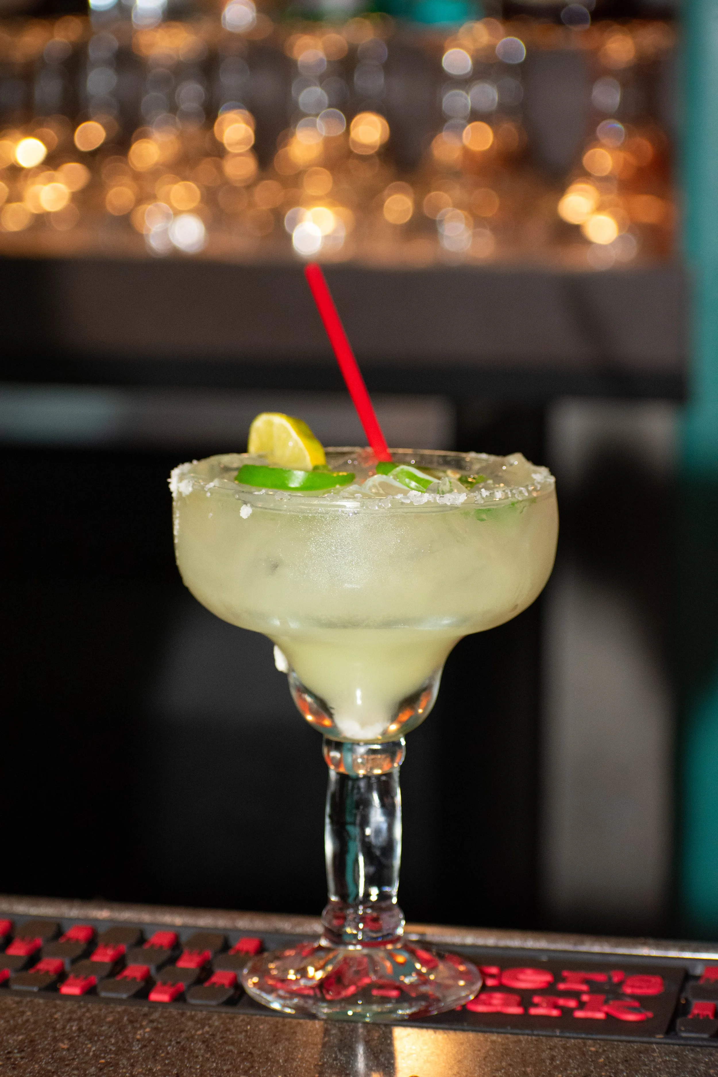 A margarita cocktail with salt on the rim, garnished with lime and jalapeño slices, and a red straw, placed on a bar counter.