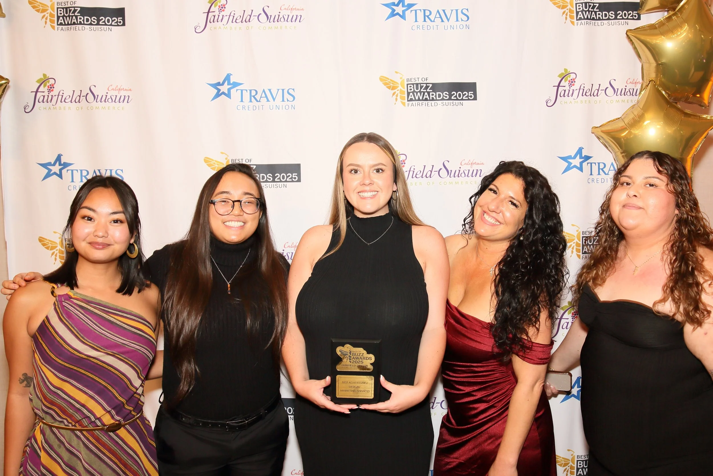 A group of six women standing in front of a backdrop with logos for the Fairfield-Suisun Chamber of Commerce, Travis Credit Union, and the Best of Buzz Awards 2025. The woman in the center is holding a trophy and is smiling.