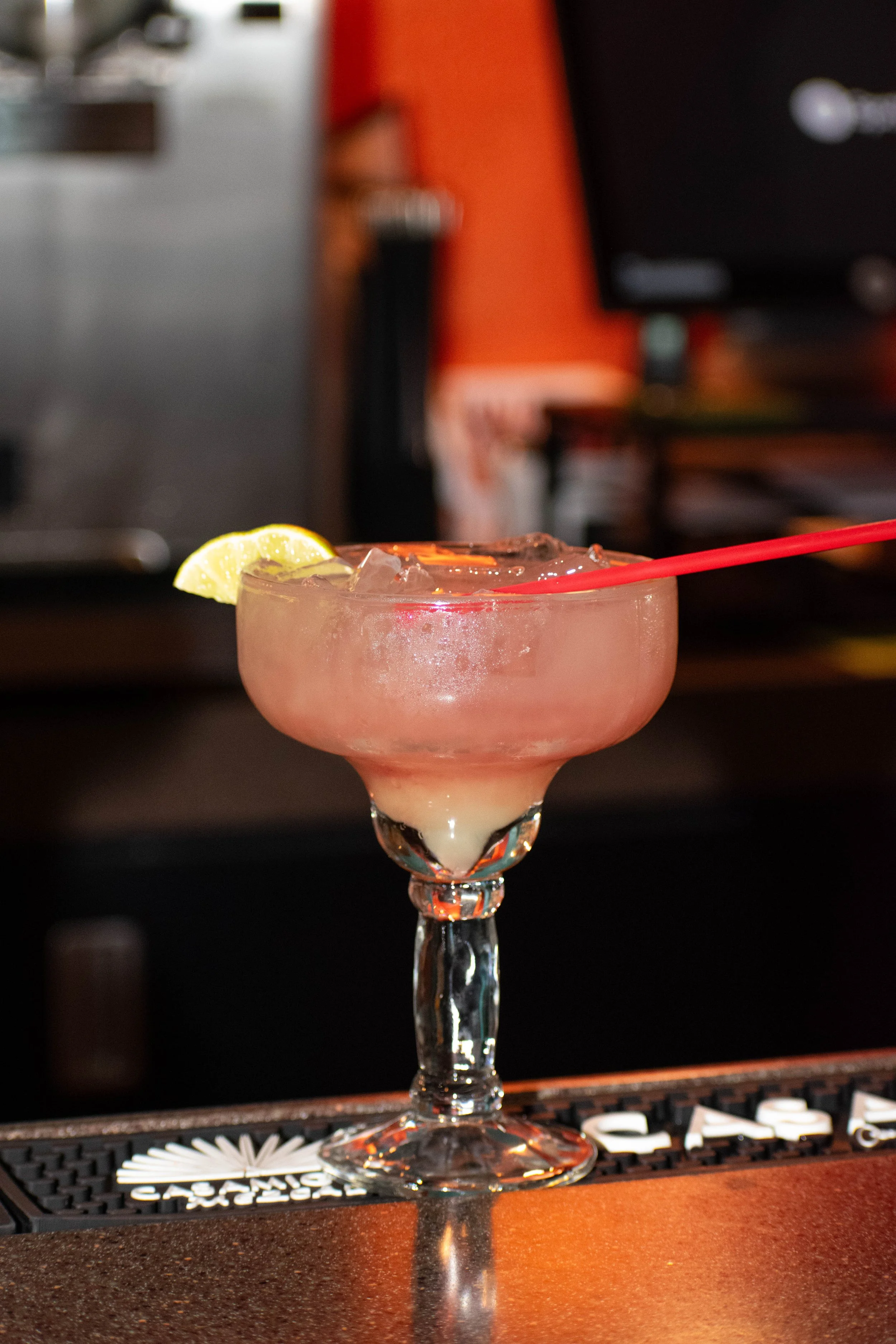 A pink cocktail with ice and a lemon wedge in a stemmed glass on a bar counter.