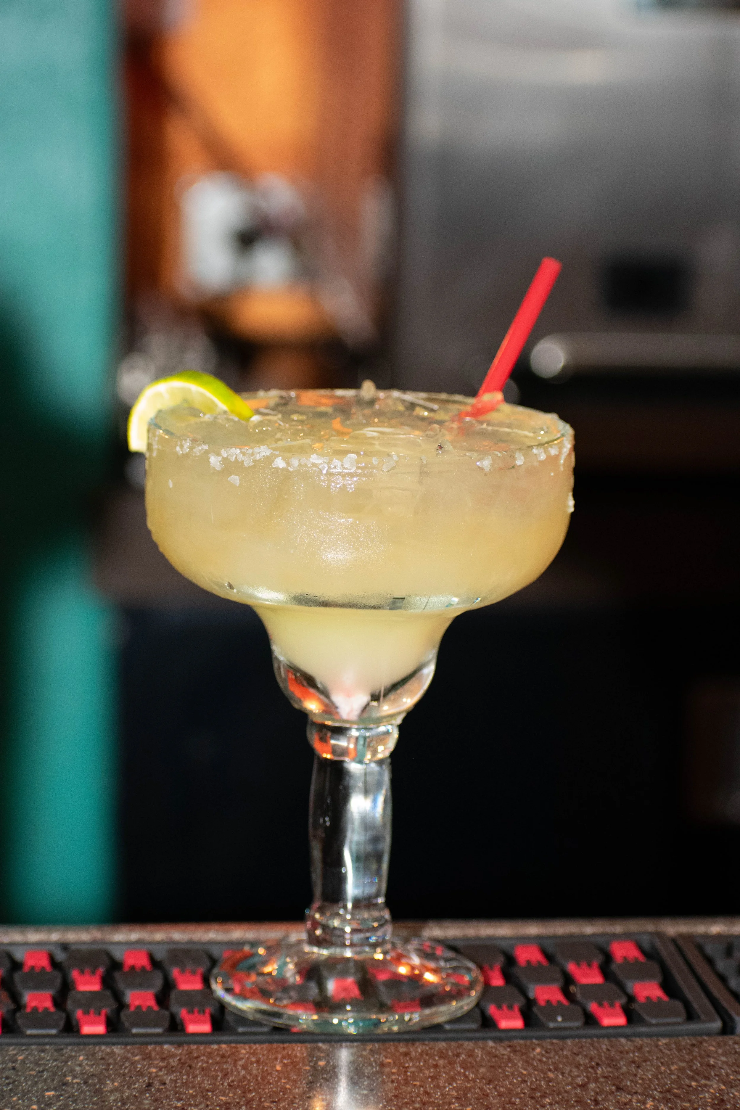 A margarita cocktail with a lime wedge and salted rim in a glass on a bar counter, with a blurred background that includes a keyboard and other bar objects.