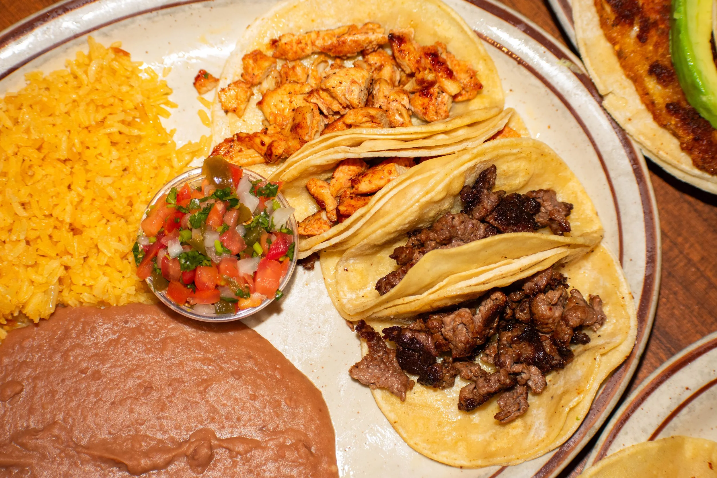 Plate of Mexican food with rice, refried beans, chopped tomato and onion salsa, and various meats inside yellow corn tacos, with another taco topped with green chili and cheese.