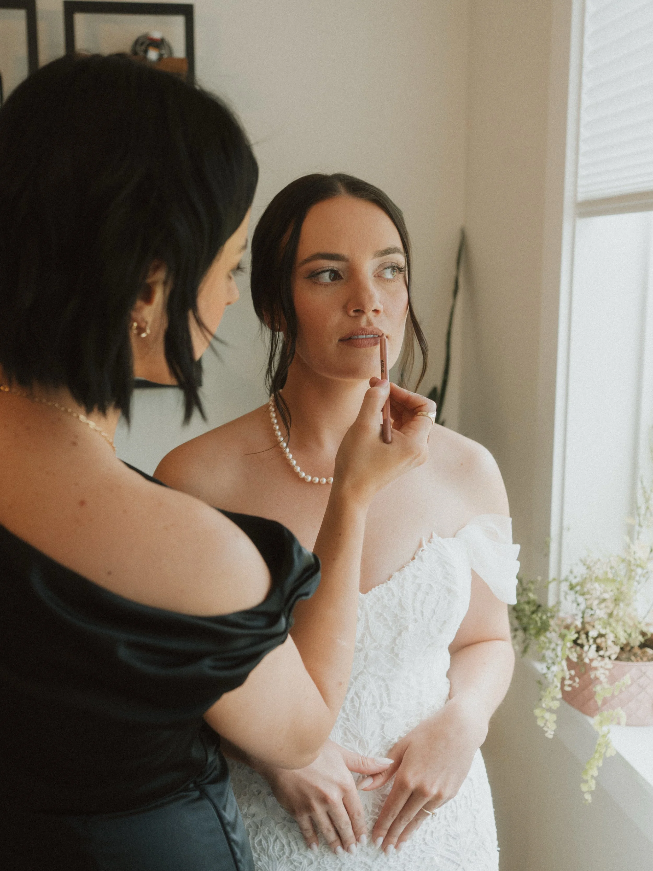 A woman in a white wedding dress is having her makeup applied by a makeup artist. She is sitting with her hands on her lap in front of a window with blinds, and floral arrangements are visible on the side.