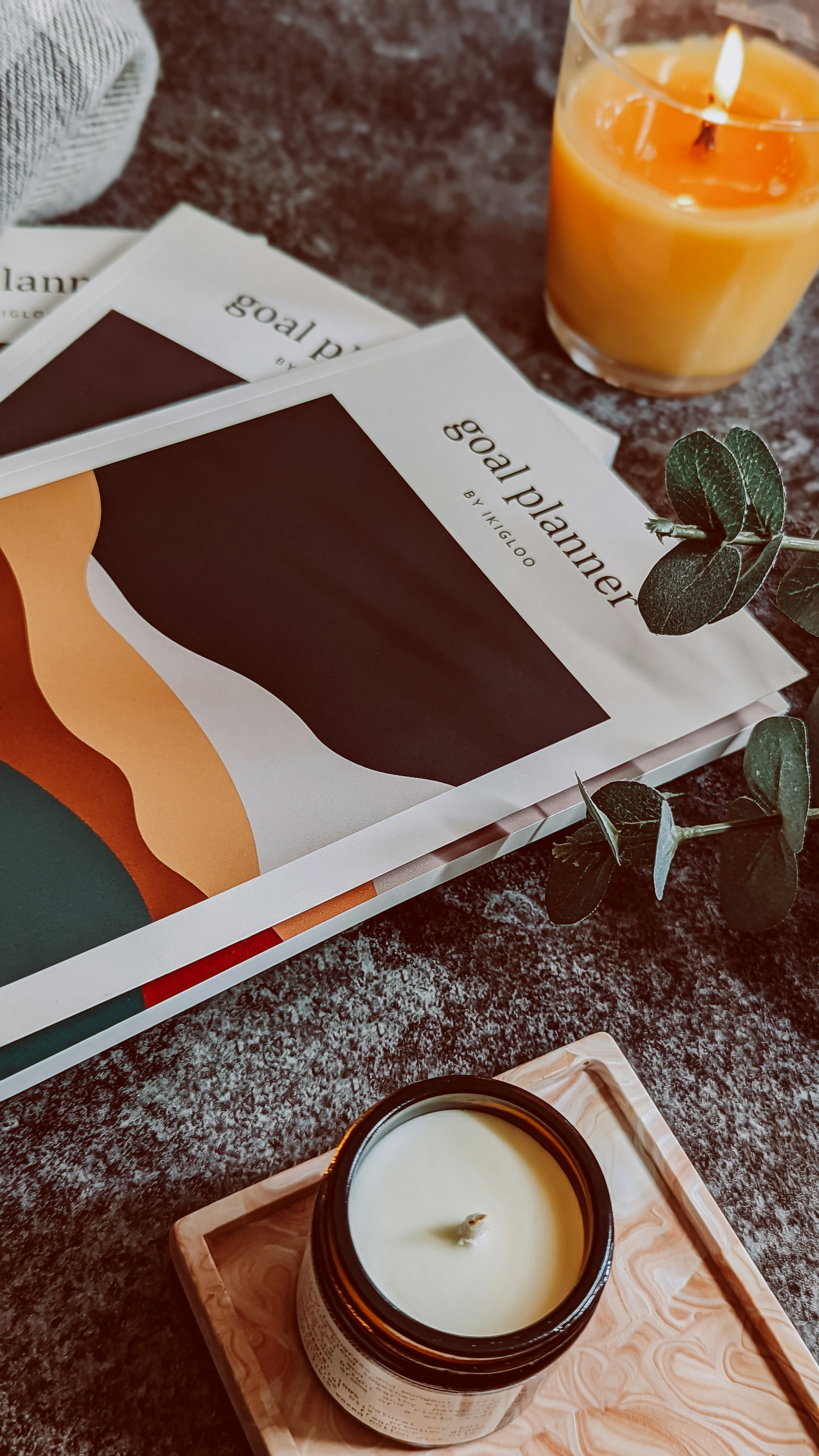 A lit candle in a dark candle jar placed on a wooden tray, with a green leafy plant next to it. In the background, there are magazines or books titled 'goal planner' and a partially visible booklet. The surface appears to be a dark, textured stone or countertop.