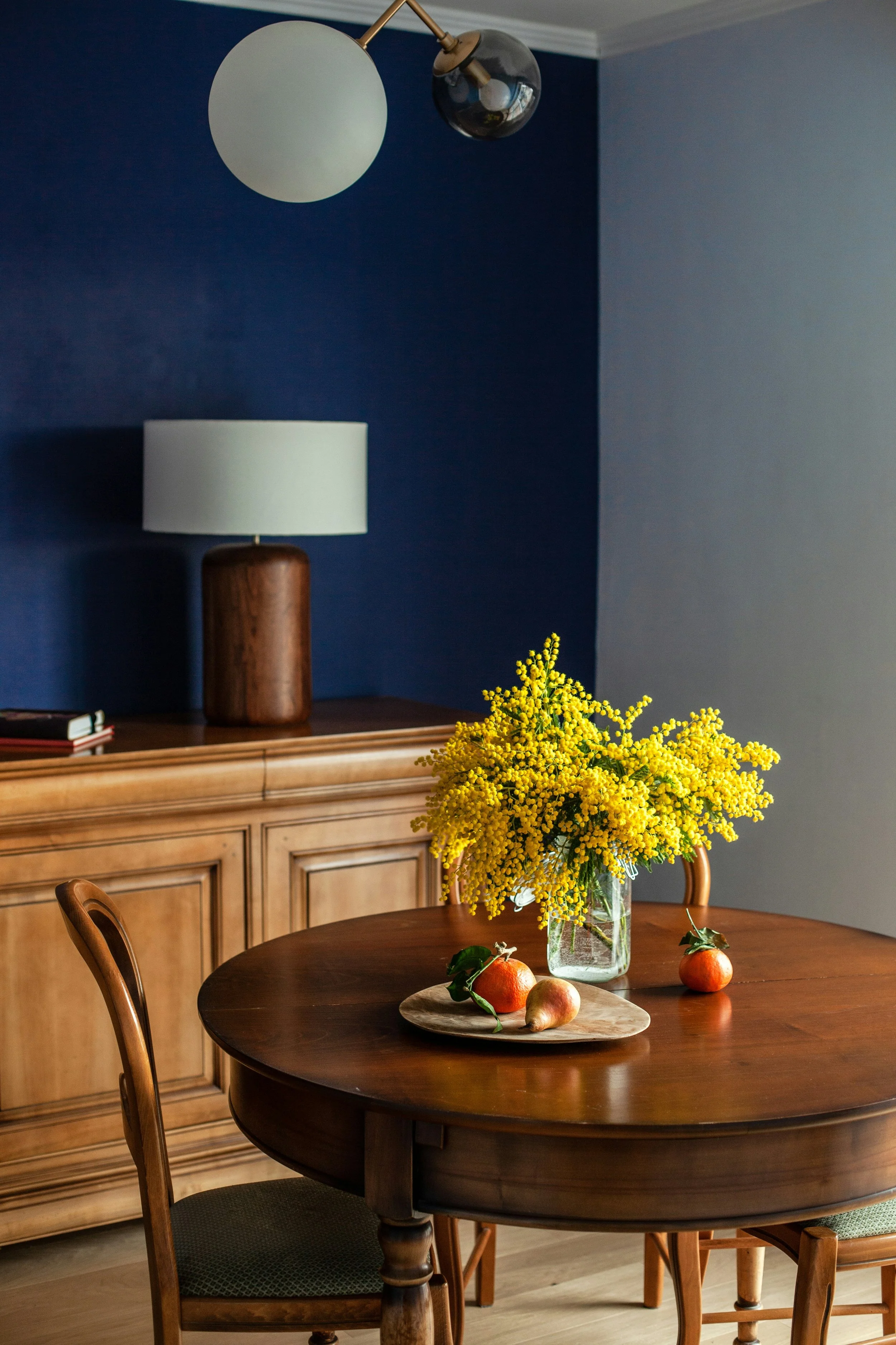 A dining room with a dark wooden round table and four matching chairs, decorated with a vase of yellow flowers and a small platter with three different fruits on it. In the background, there is a wooden sideboard and a wall painted in a dark teal color with white crown molding. A modern table lamp with a white shade and a wooden base is on the sideboard, and a ceiling light fixture with a white and black globe hangs from the ceiling.