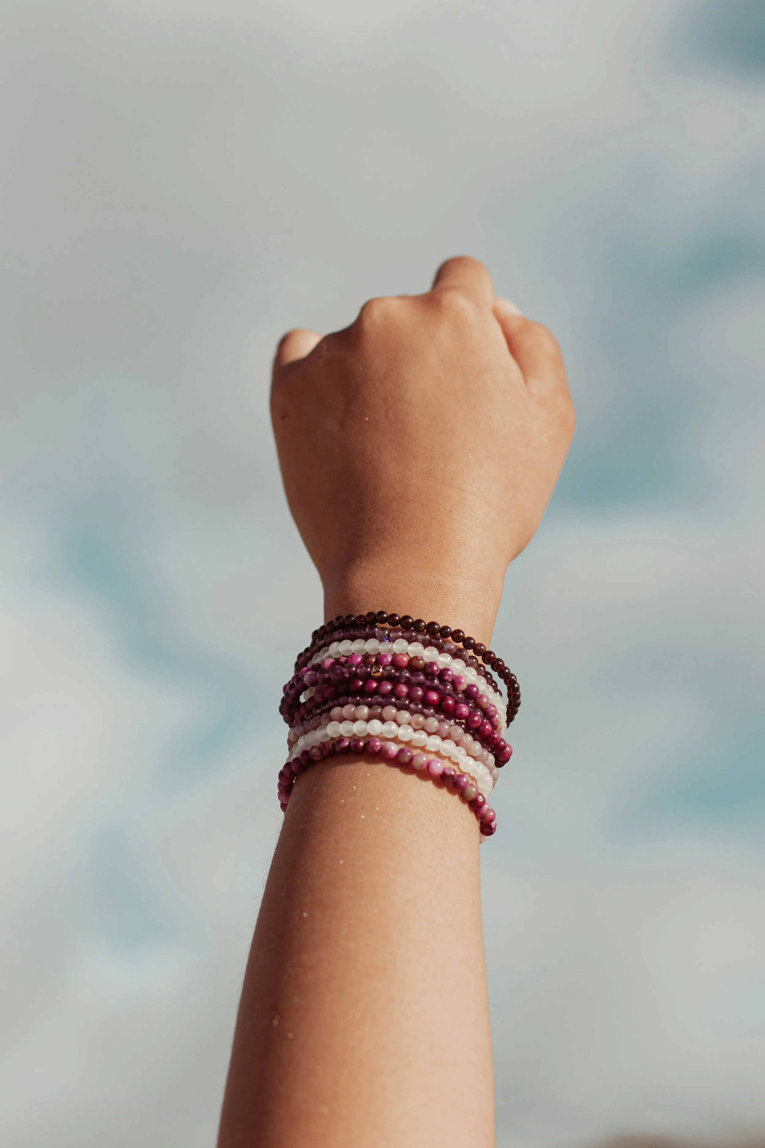 A person's hand raised upward with multiple colorful beaded bracelets on the wrist, set against a cloudy sky background.