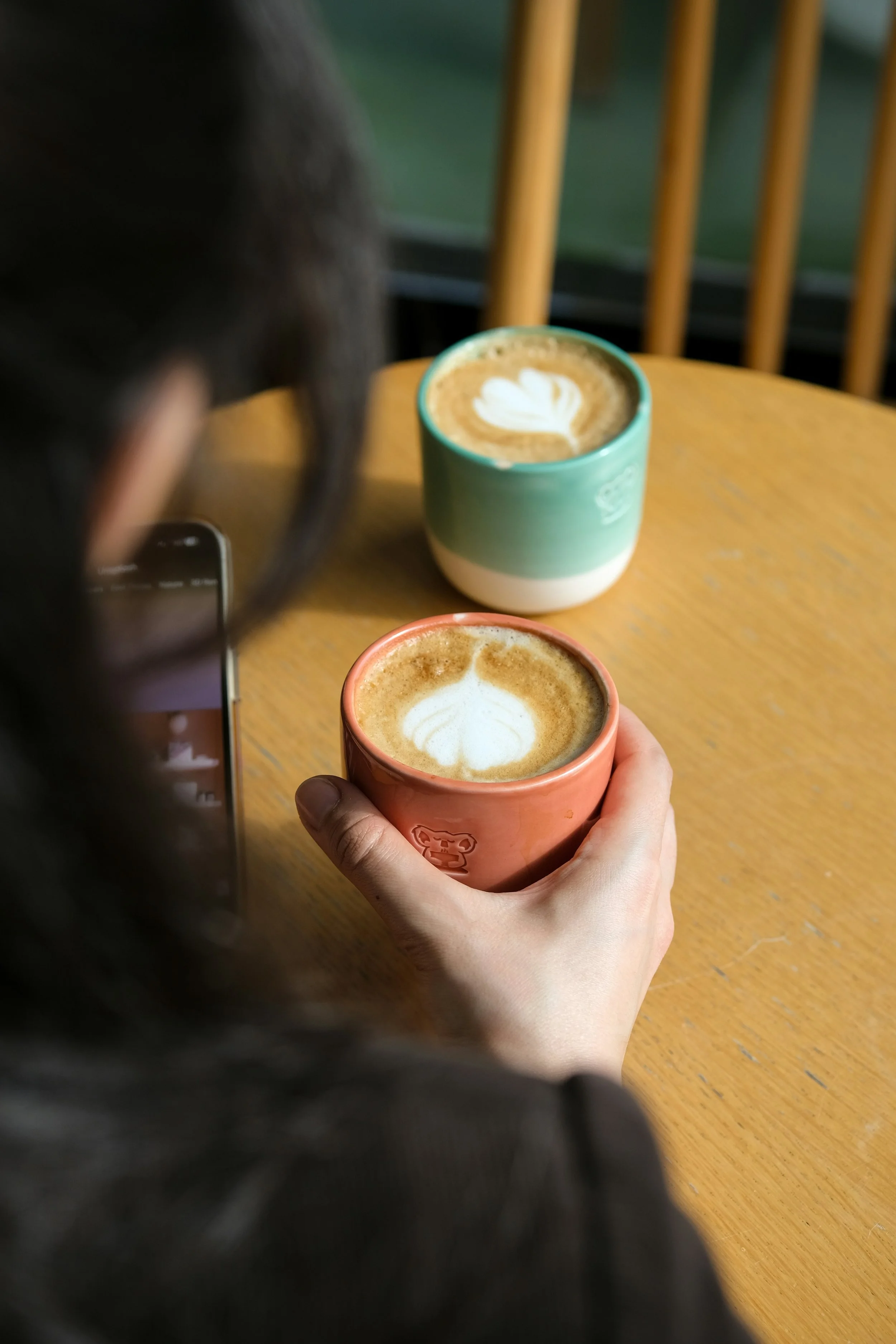 A person holding a pink ceramic cup of latte with a leaf-shaped foam design, with another person in the background holding a teal cup of latte with a similar foam design, both on a wooden table.