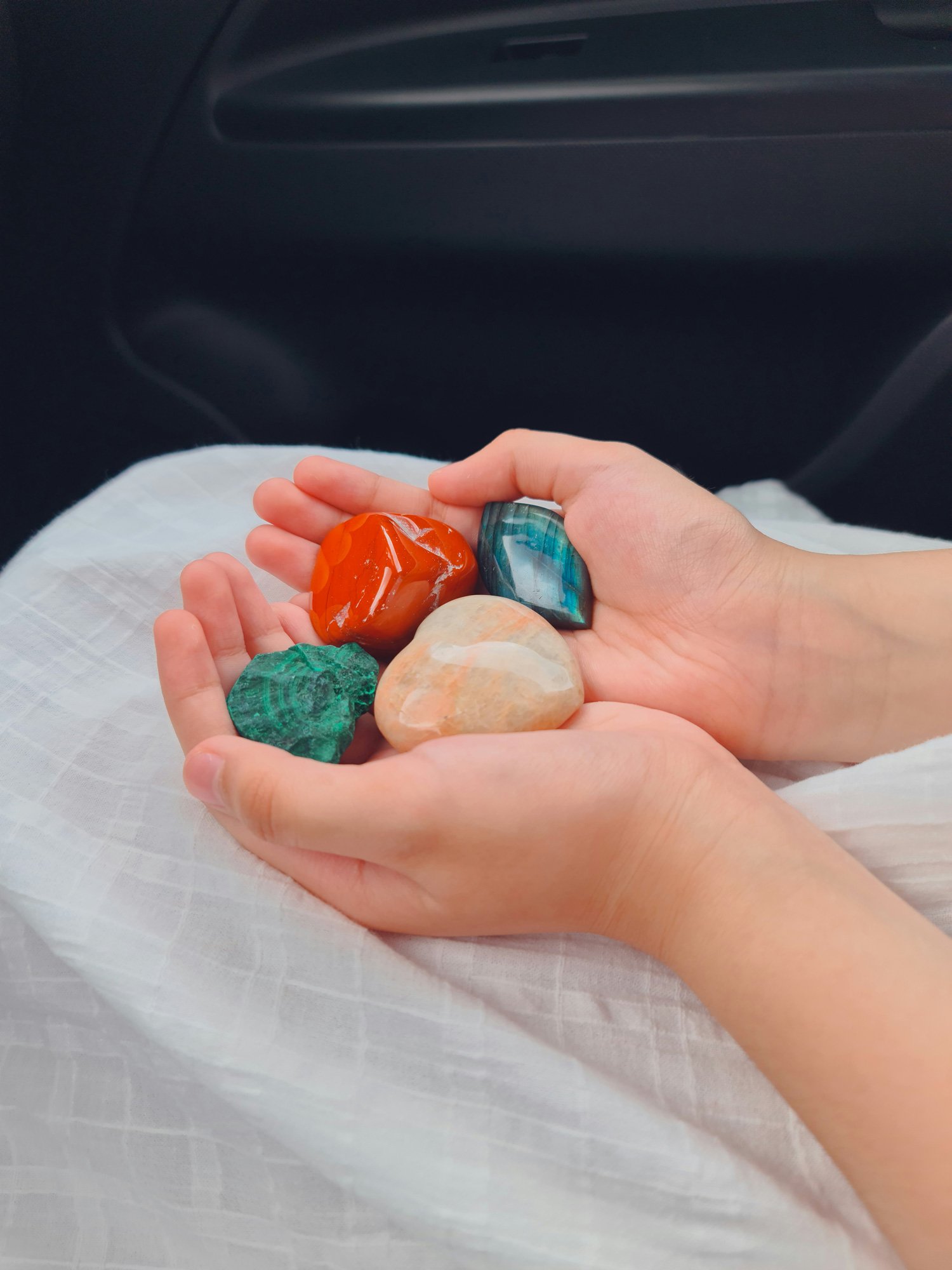 A person holding several polished stones of various shapes and colors, including orange, green, blue, and beige, in their hands.