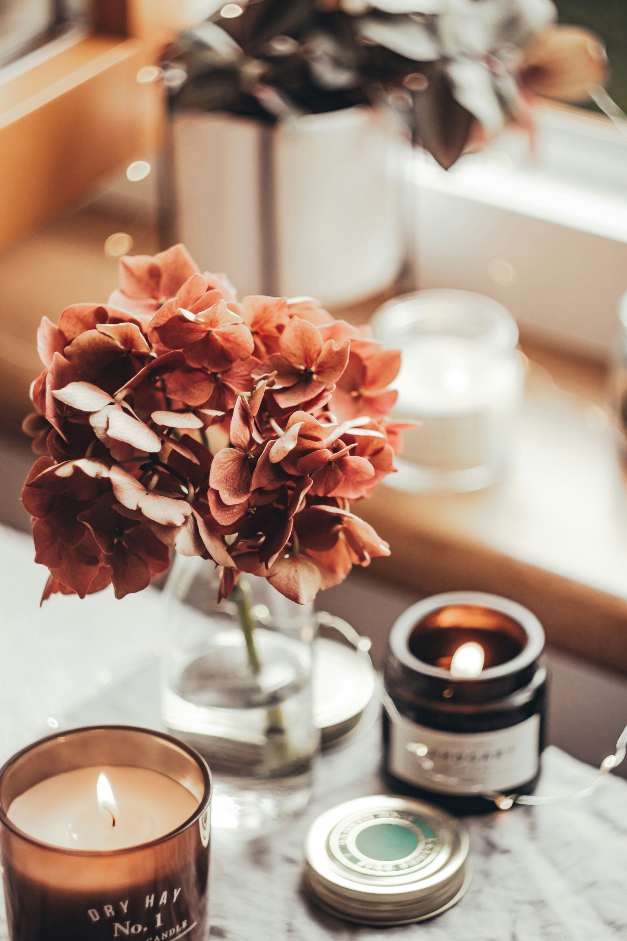 A dried pink hydrangea flower in a glass vase, surrounded by lit candles in candle holders, with a blurred background of potted plant and decorative items on a tray.