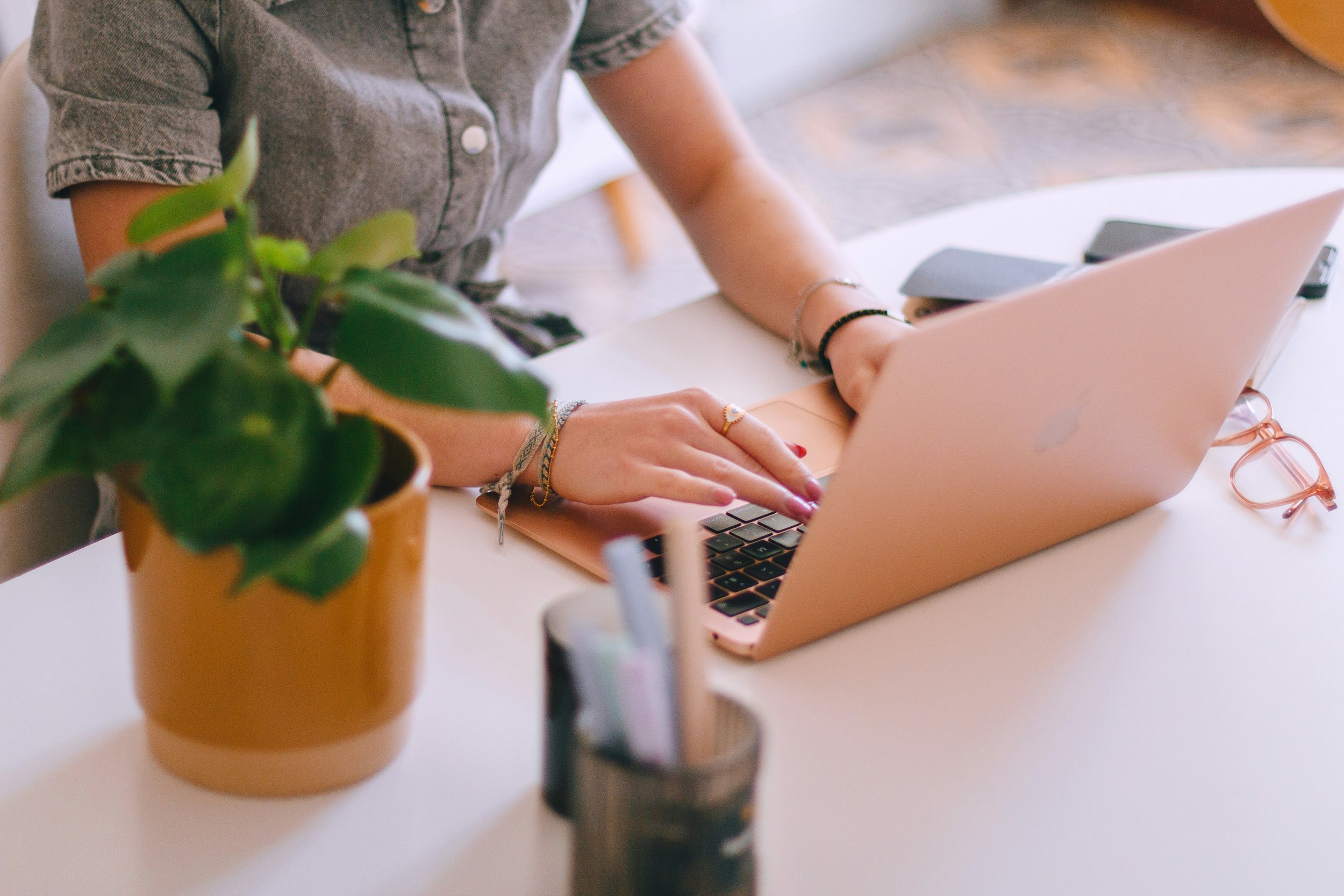 Person working on a laptop at a white desk with a potted plant in the foreground and a smartphone nearby.