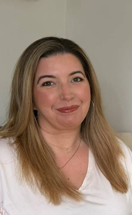 A woman with medium-length blonde hair, smiling, wearing a white top and gold hoop earrings.