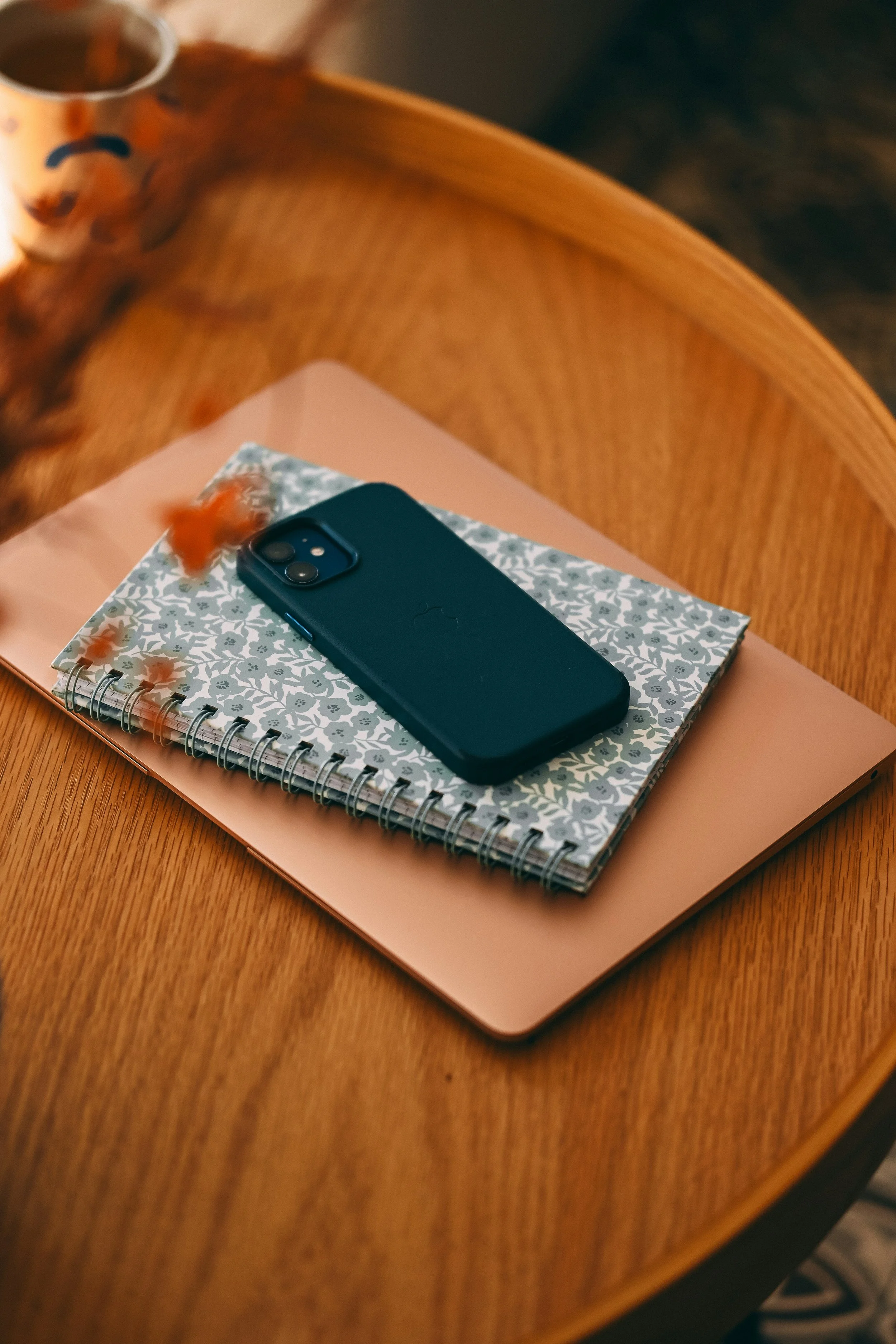 A blue smartphone resting on a floral-patterned notebook placed on top of a pink folder, all on a wooden table.