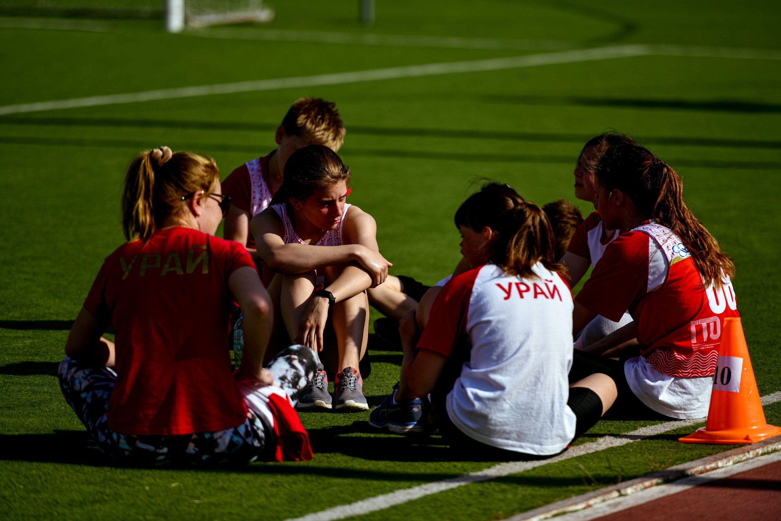 A group of young female athletes in red and white uniforms sitting on a sports field, listening to a coach during a training session.