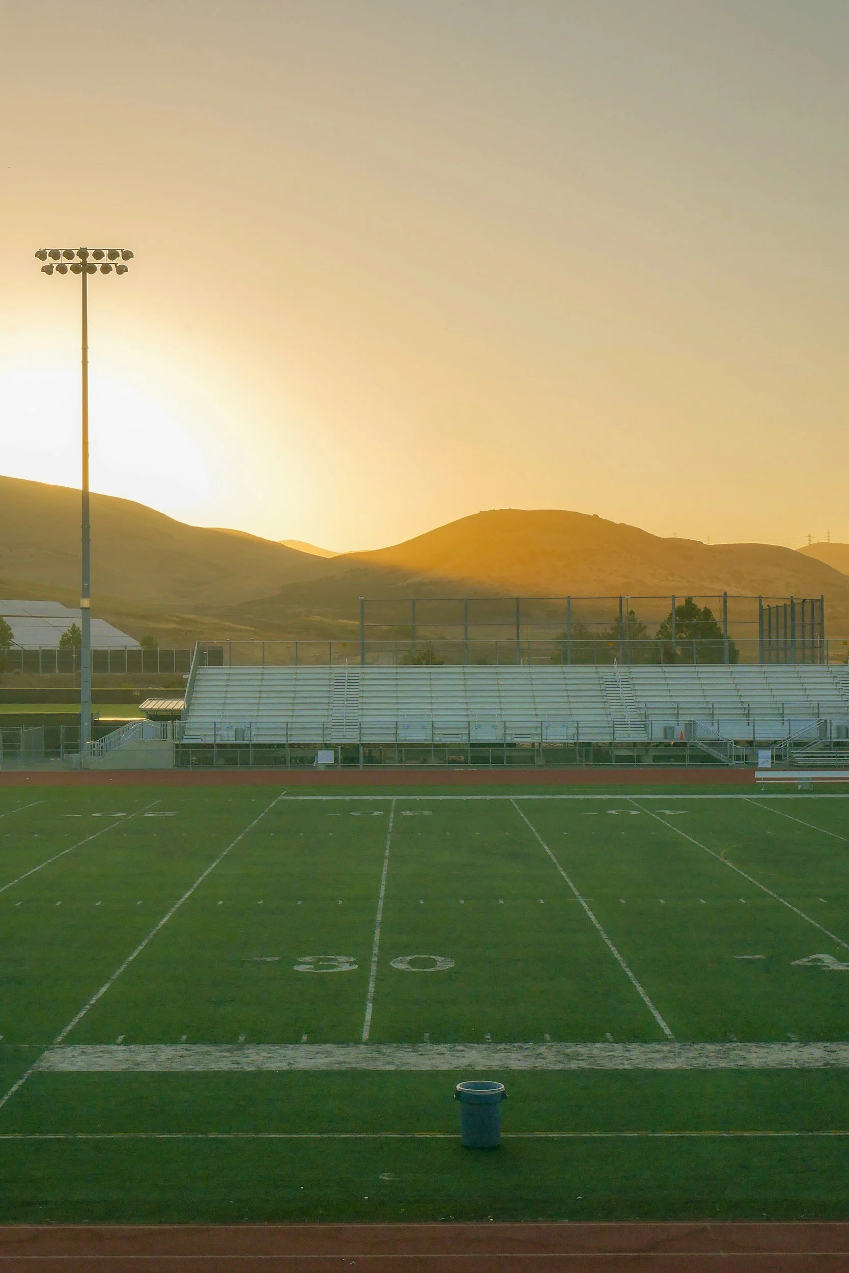 An empty outdoor football field during sunset with stadium bleachers, a goalpost with stadium lights, a trash can, and mountains in the background.