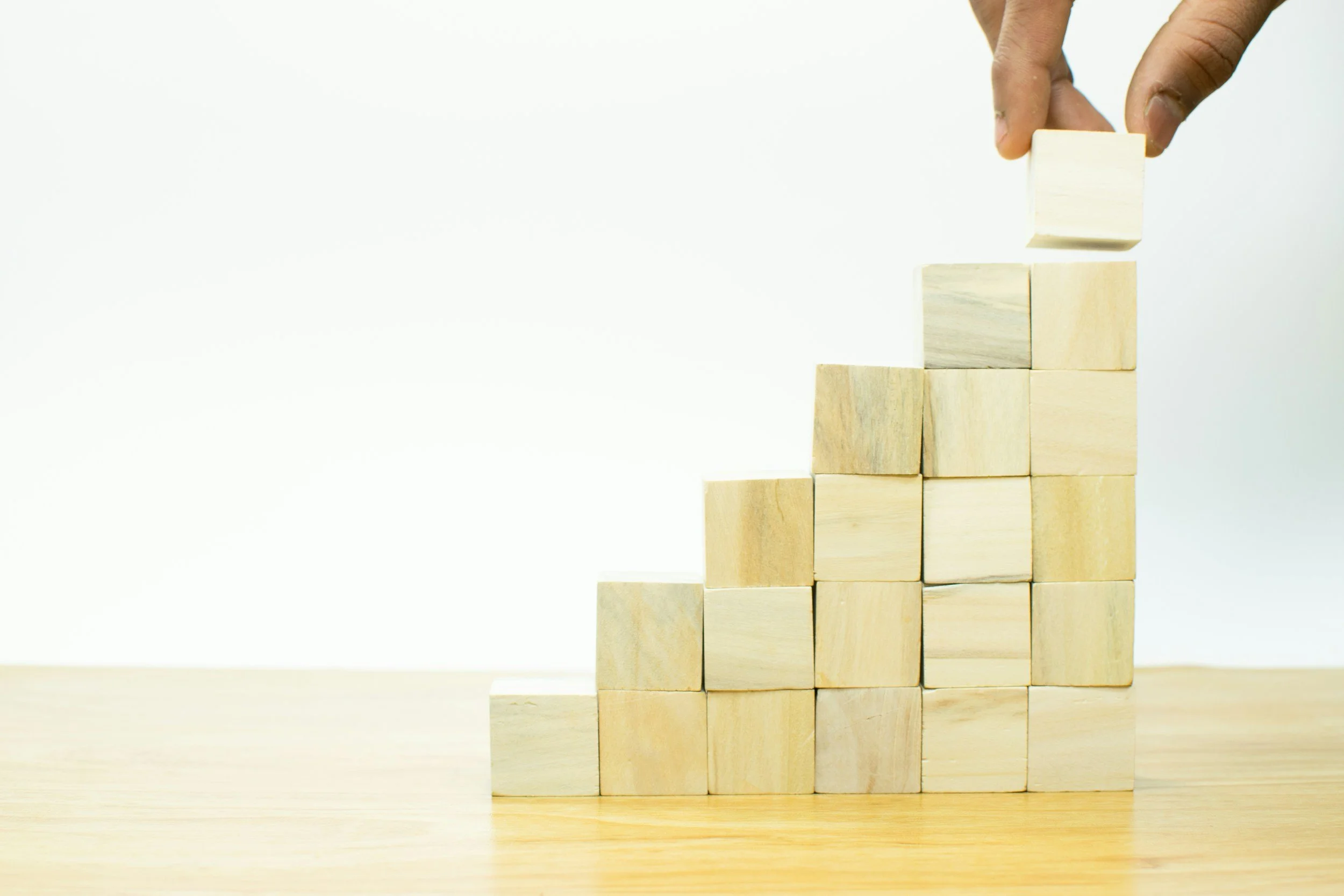 A hand placing a small wooden block on top of a pyramid of other wooden blocks, with the blocks stacked in a staircase pattern.