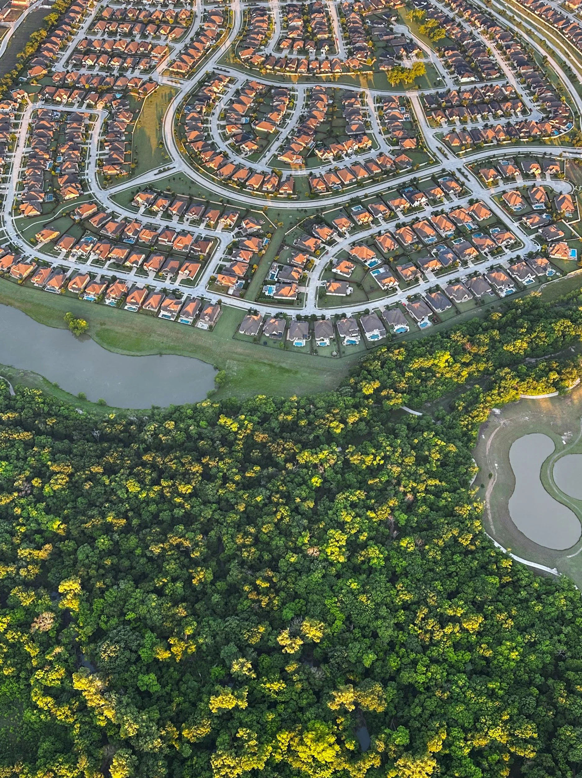 Aerial view of a residential neighborhood with many houses featuring orange roofs. The neighborhood is arranged in a circular pattern with multiple curved streets. Surrounding the houses are green lawns and a body of water with dense trees at the bottom of the image.
