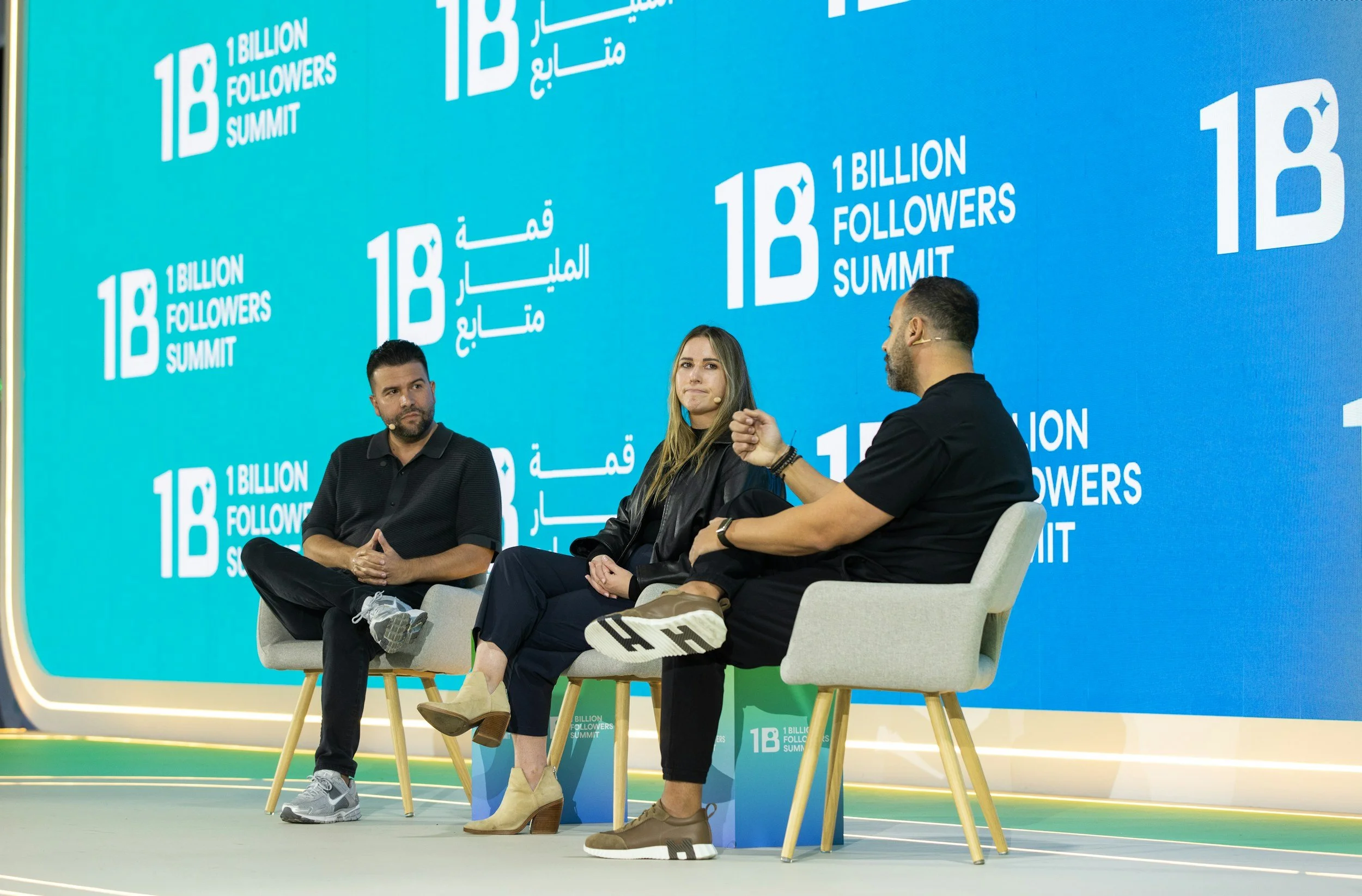 Three people sitting on stage in front of a blue backdrop with the event name '18th Billion Followers Summit' in English and Arabic. They are engaged in conversation, with one man gesturing as he speaks. The person on the left has dark hair and beard, wearing a black shirt and gray sneakers. The woman in the middle has long hair, beige high heels, and is wearing a black jacket. The man on the right has dark hair and beard, wearing a black shirt, dark pants, and white sneakers.