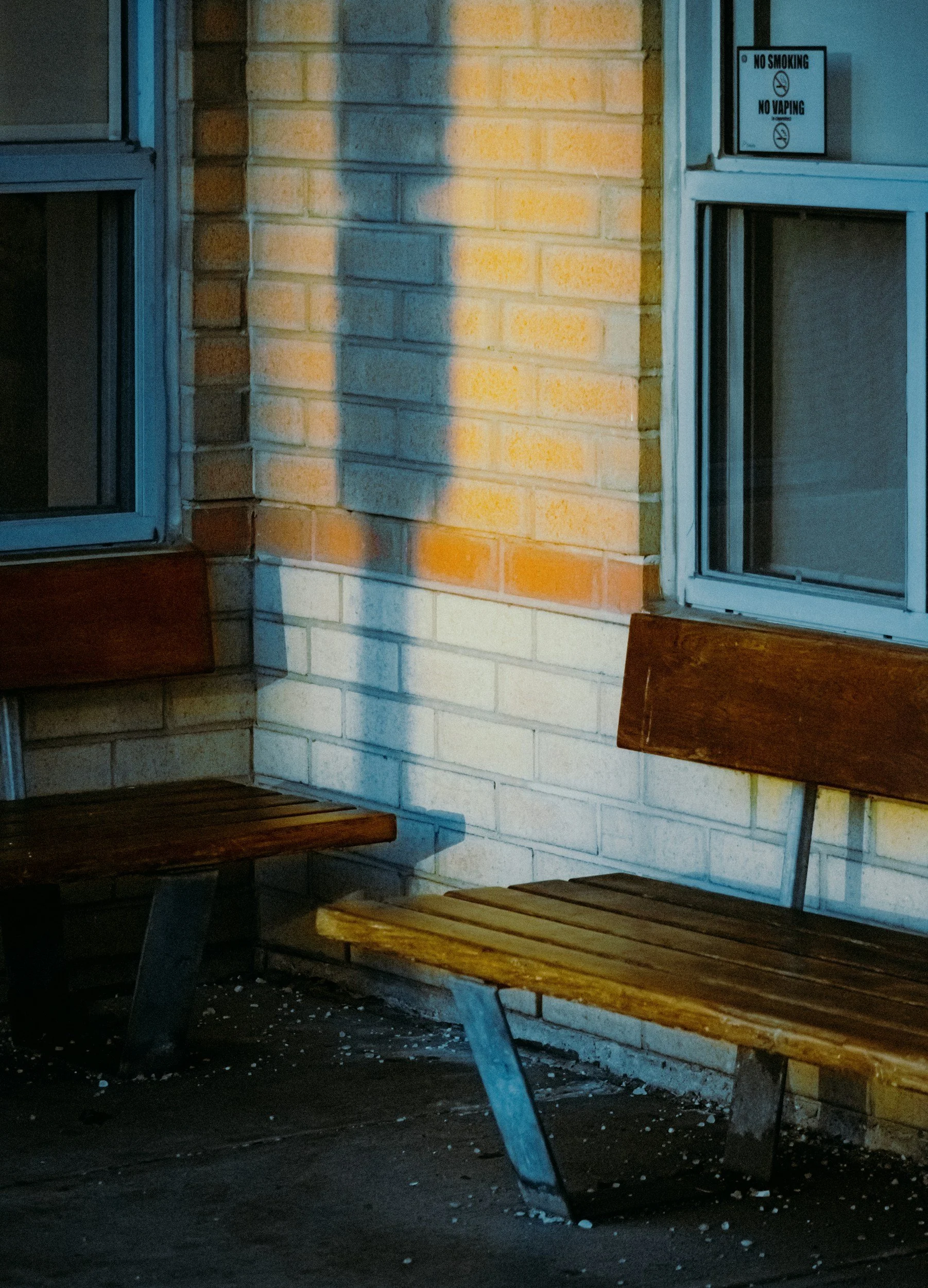 Exterior view of a brick wall with two wooden benches and a window, illuminated by a yellow streetlight. There is a sign on the wall that says 'No Smoking, No Vaping'.